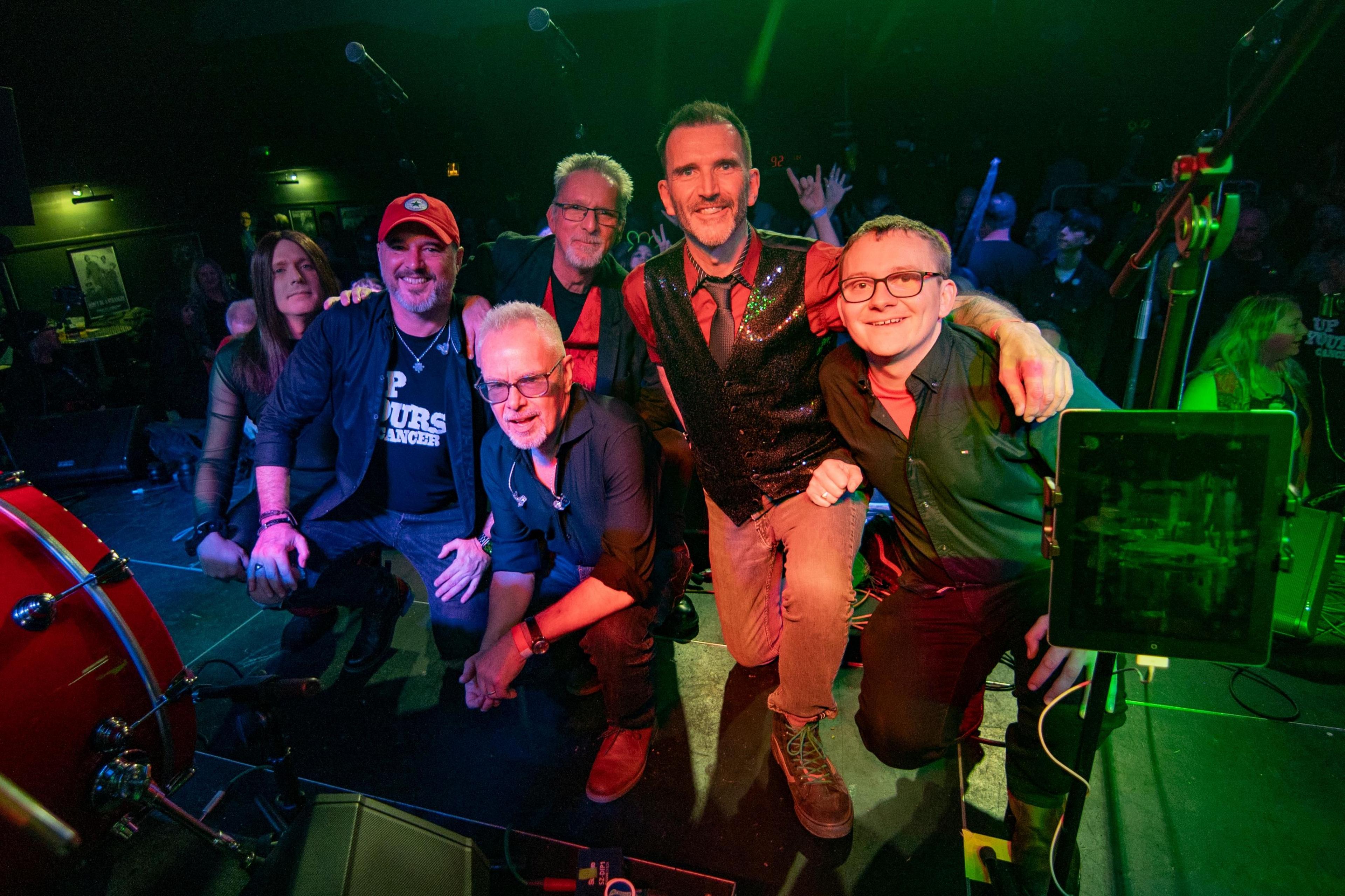 A group of six musicians with their backs to the stage. They are crouched down next to a drum. People can be seen in the background.