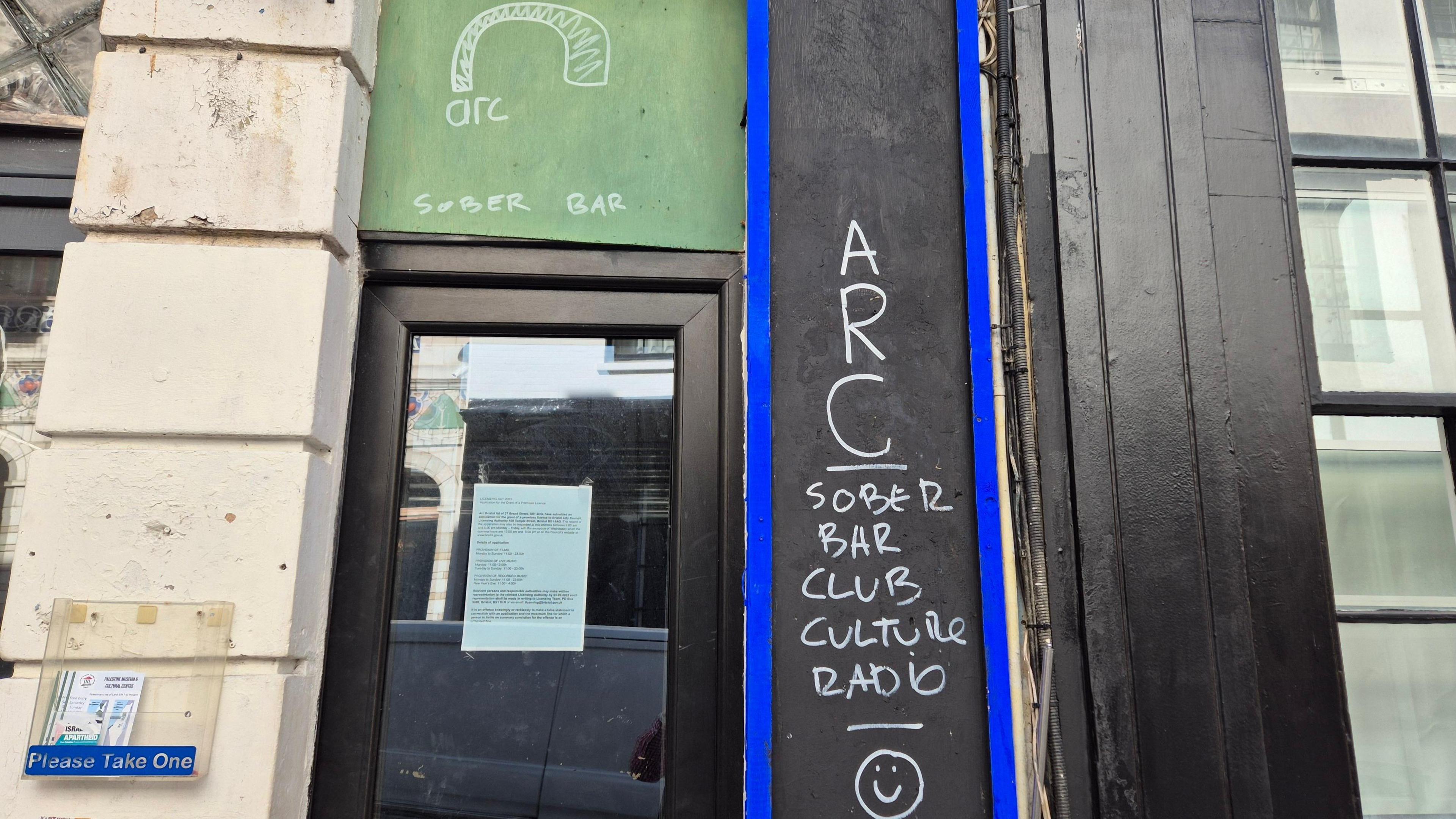 A door to the Arc bar. It has a glass window on it that has a planning permission letter taped to it. Above it someone has written in chalk "Arc - Sober Bar" and to the right on a slim chalk board someone has written "ARC - Sober bar, club, culture, radio" with a smiley face beneath it.
