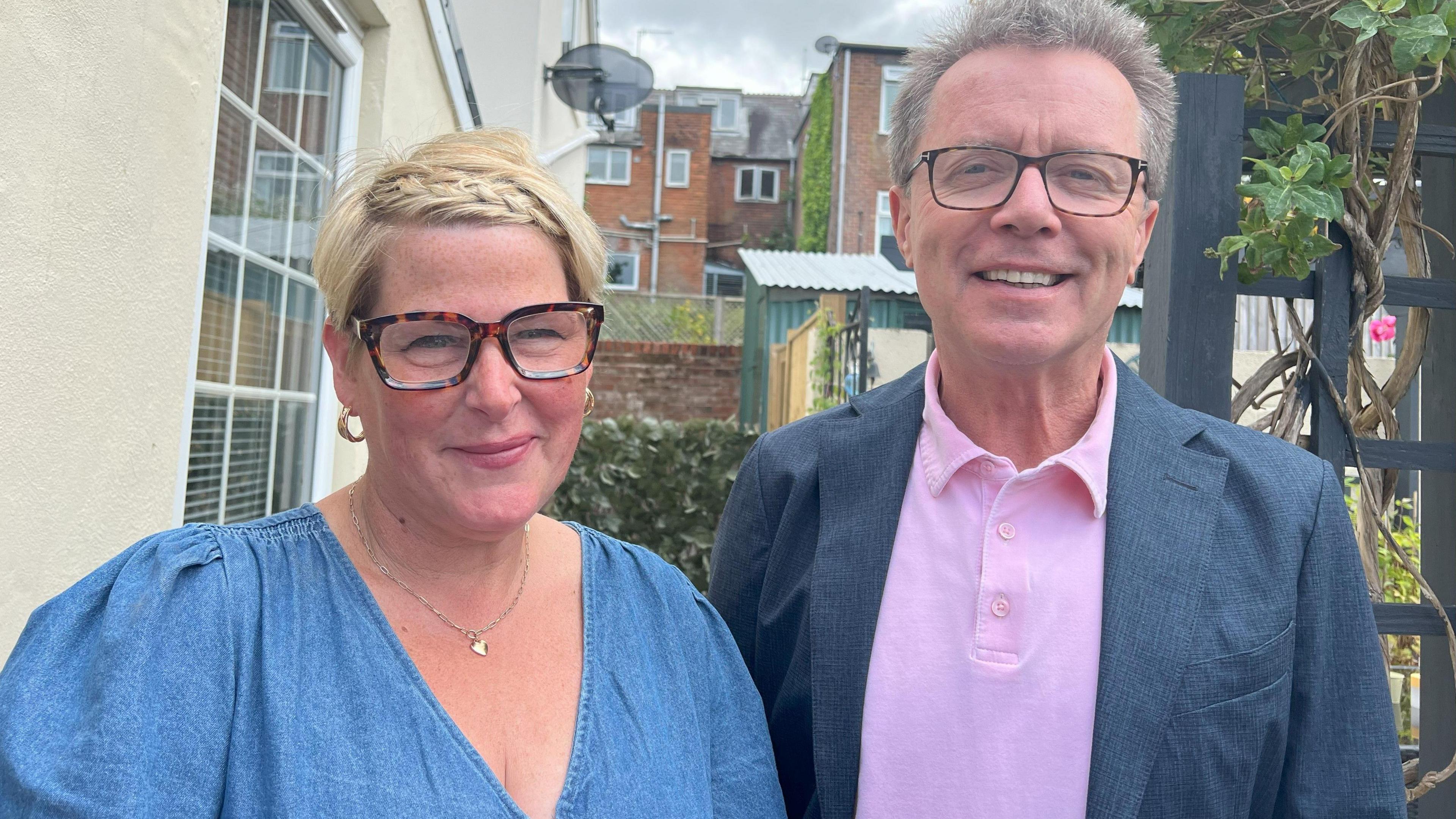 Hilary and Nicky Campbell stand together smiling at the camera. Both are wearing glasses. They stand in a garden. Hilary is wearing a blue dress, Nicky is in a suit jacket with a pink polo top underneath