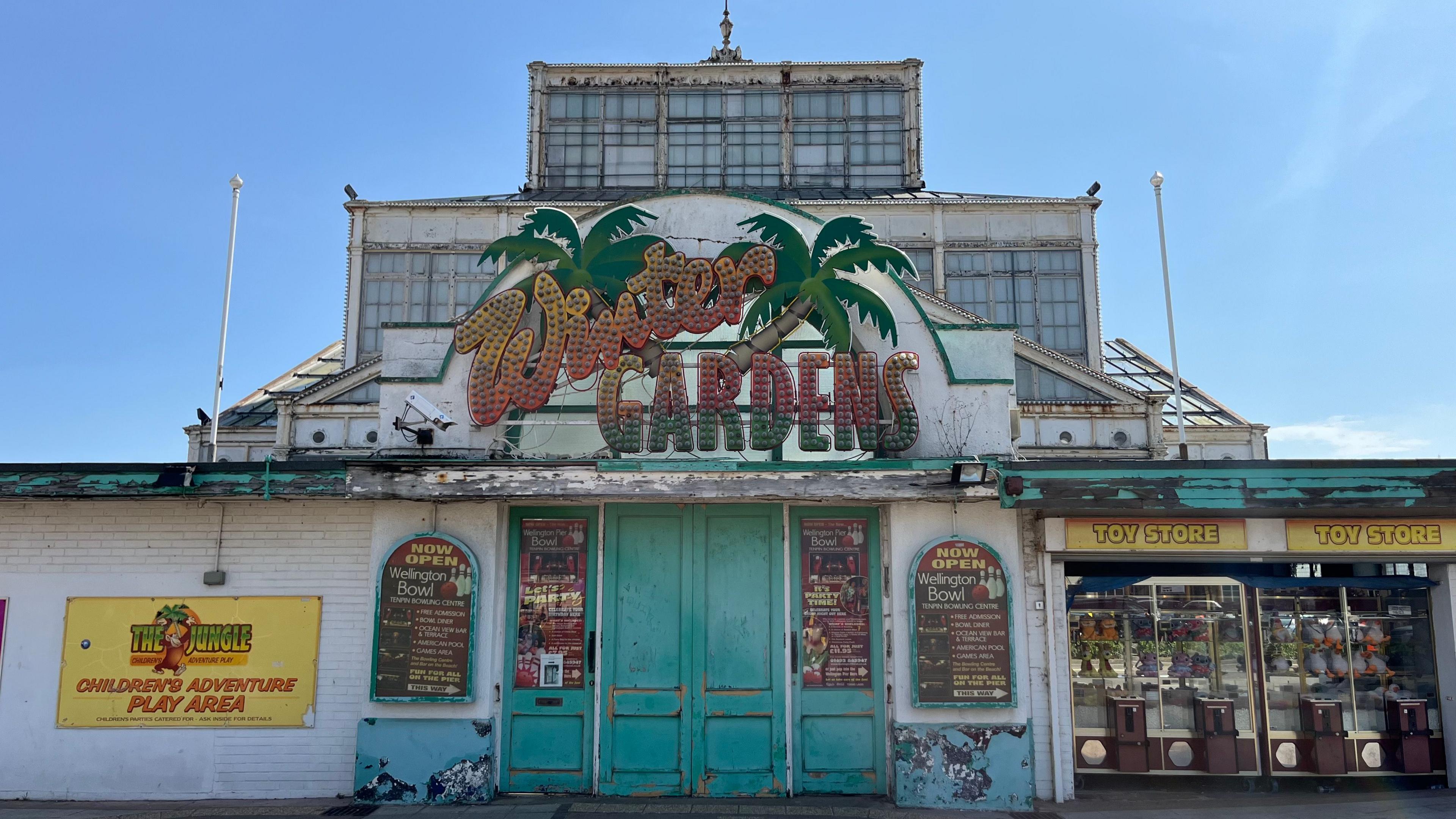 The outside of the Winter Gardens in Great Yarmouth, Norfolk. On the front of the building is a sign which previously used to illuminate. The sign which also has palm trees on it reads, Winter GARDENS. 