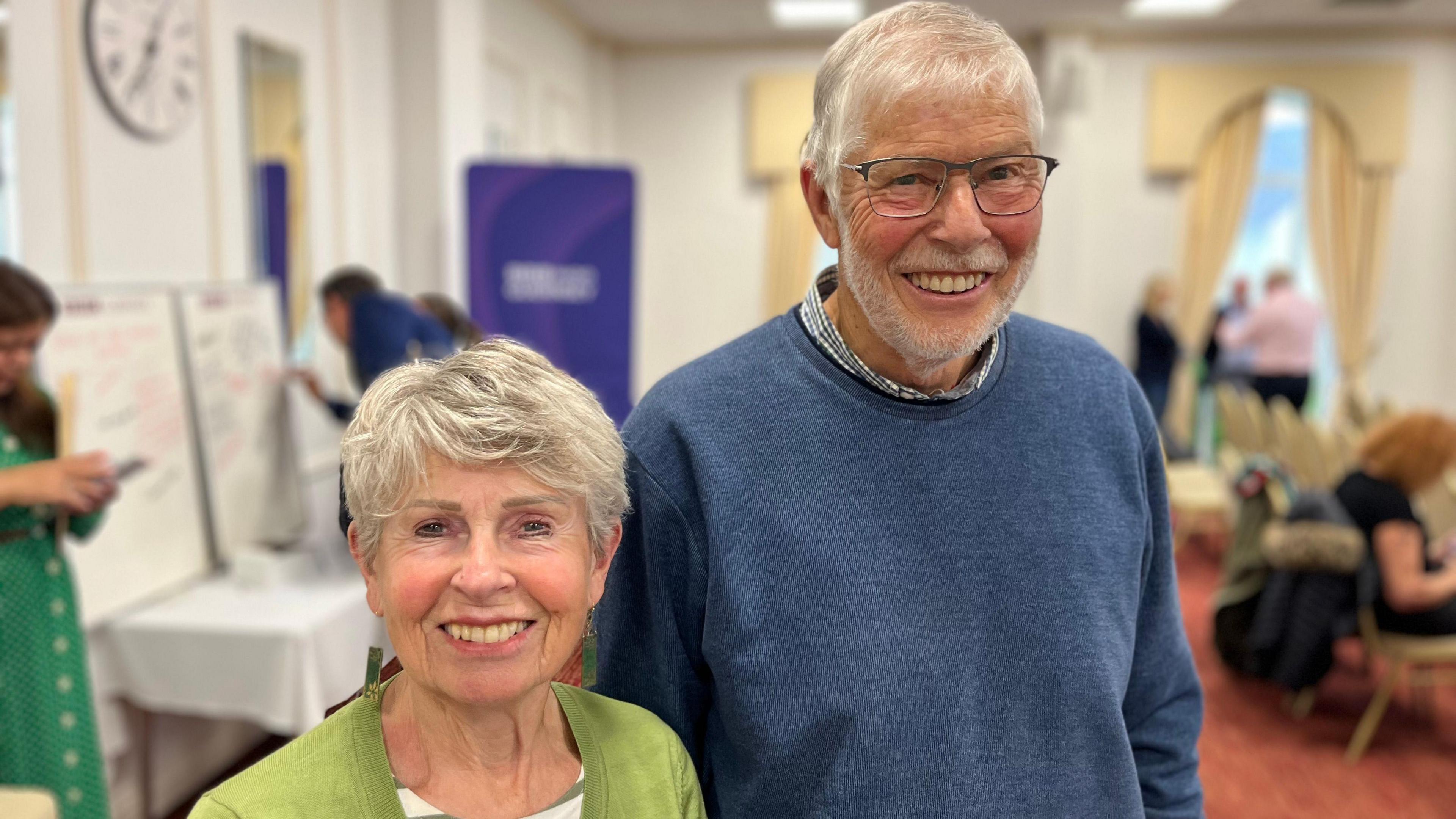 On the left is a shorter woman with short grey hair. She's wearing a green jacket. On her right is a taller man, he's wearing a blue jumper, has grey hair and is wearing blue glasses.