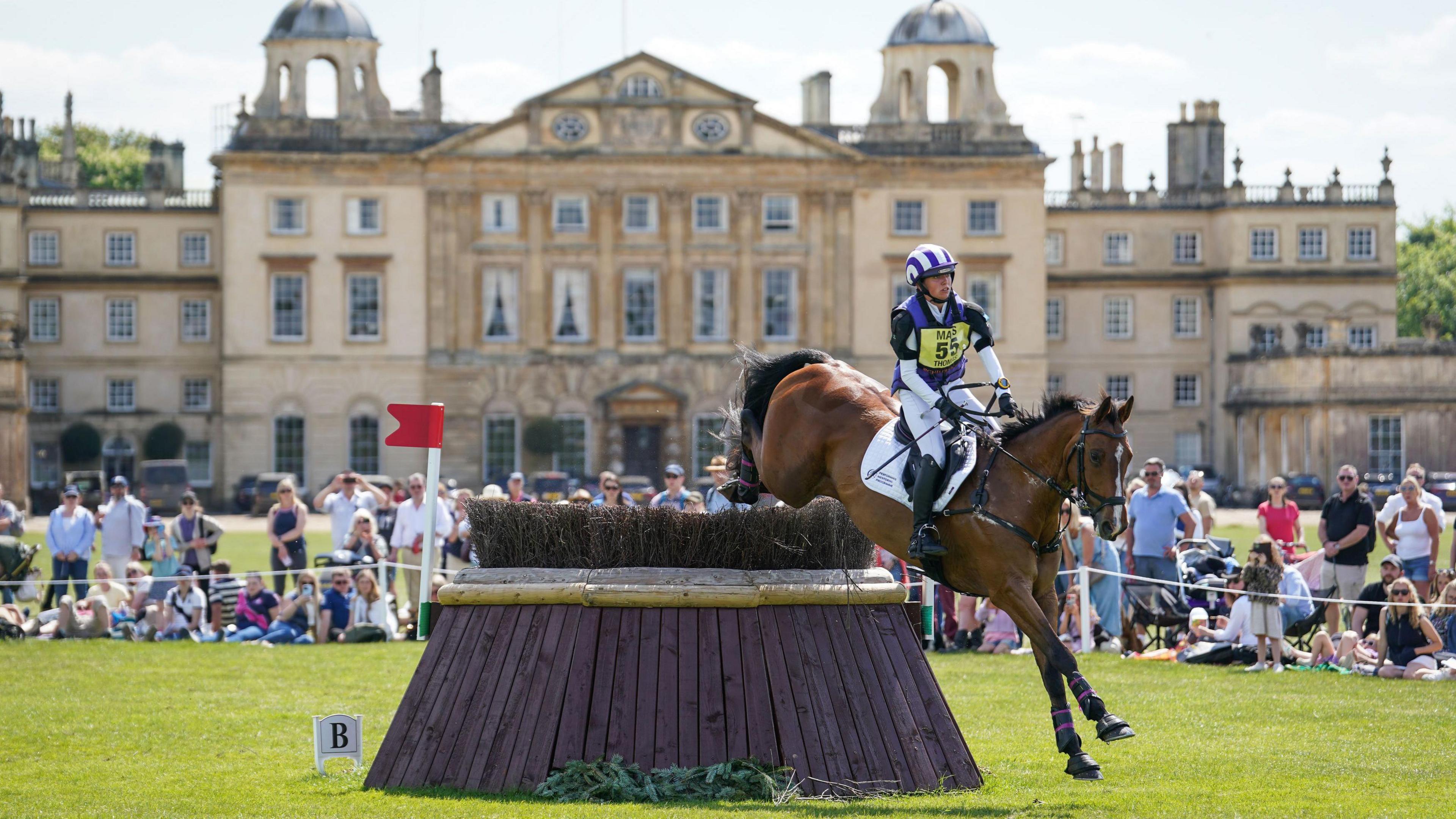 A horse and rider clear a jump obstacle in front of the main house at the Badminton Estate in Gloucestershire. In the background several spectators can be seen watching on