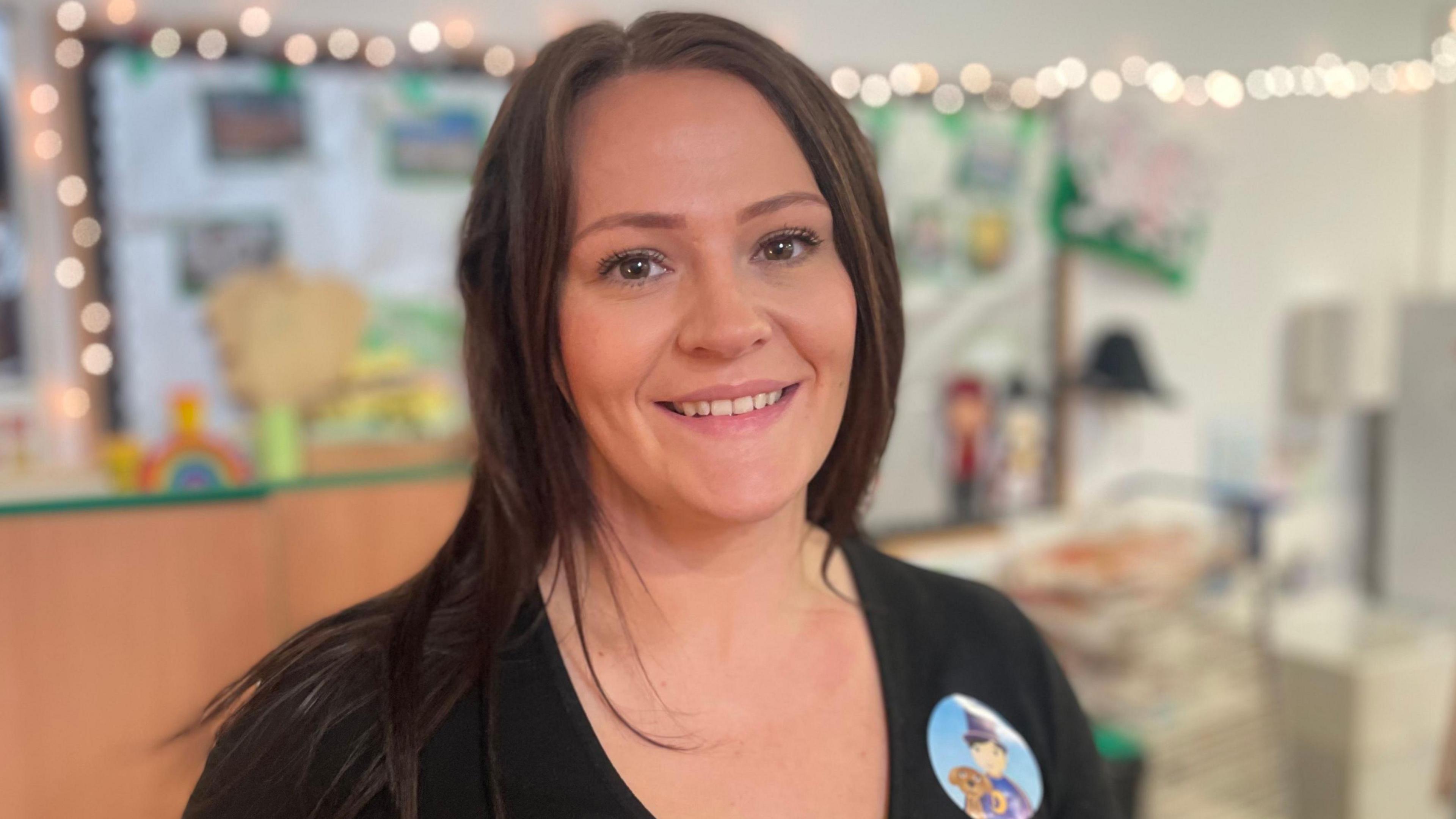 Nia-Ffion gyda gwallt hir tywyll yn gwenu, gyda goleuadau bach yn disgleirio ar wal y fethrinfa tu ol iddi.