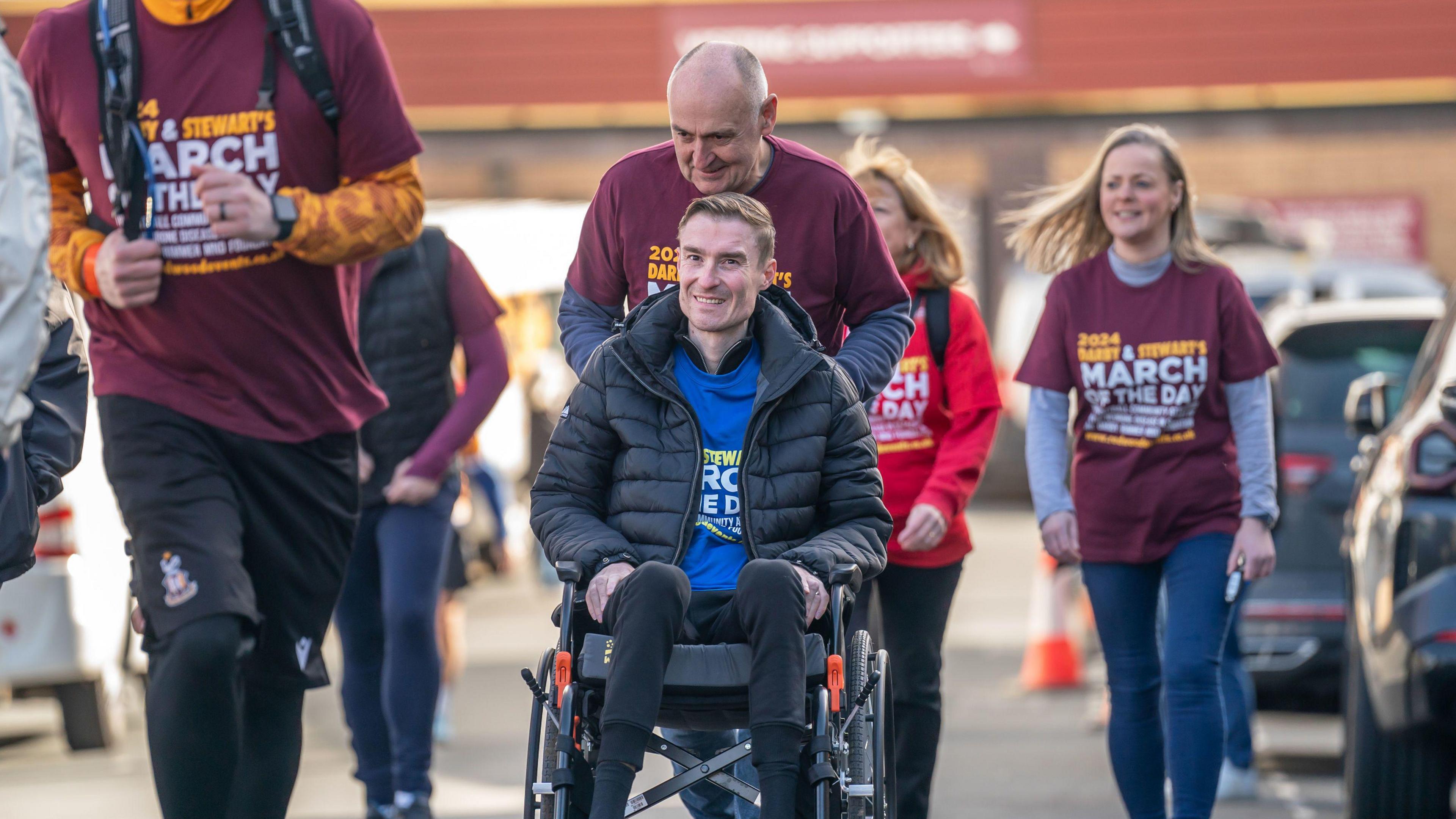 Stephen Darby is dressed in a blue "March Of The Day" t-shirt and an unzipped black coat. He smiles whilst being pushed in a wheelchair by another man dressed in a burgundy "March Of The Day" t-shirt.