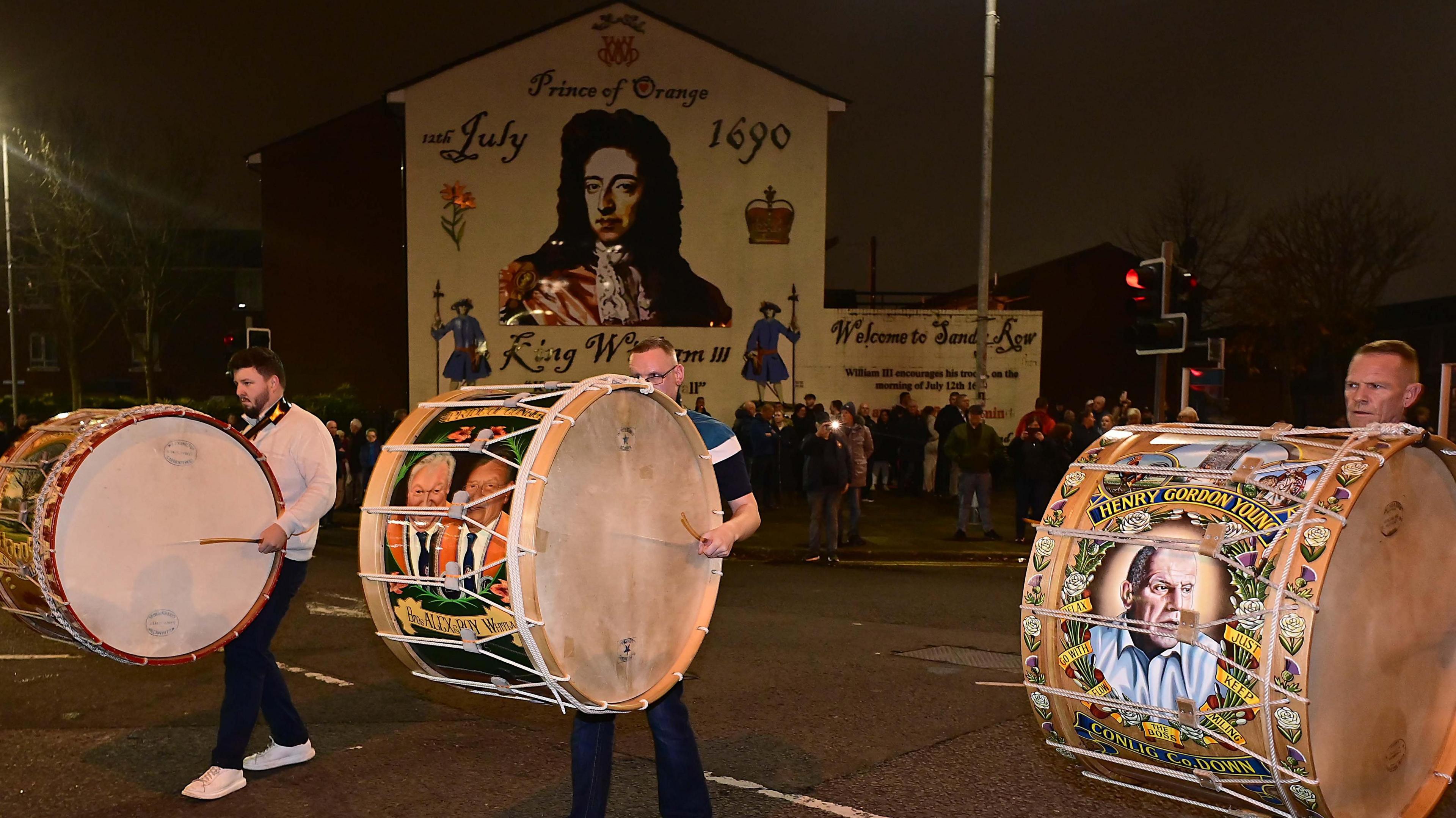 Three men are playing large lambeg drums on the road in front of a mural of King William III. It's night and a crowd is standing around the mural.