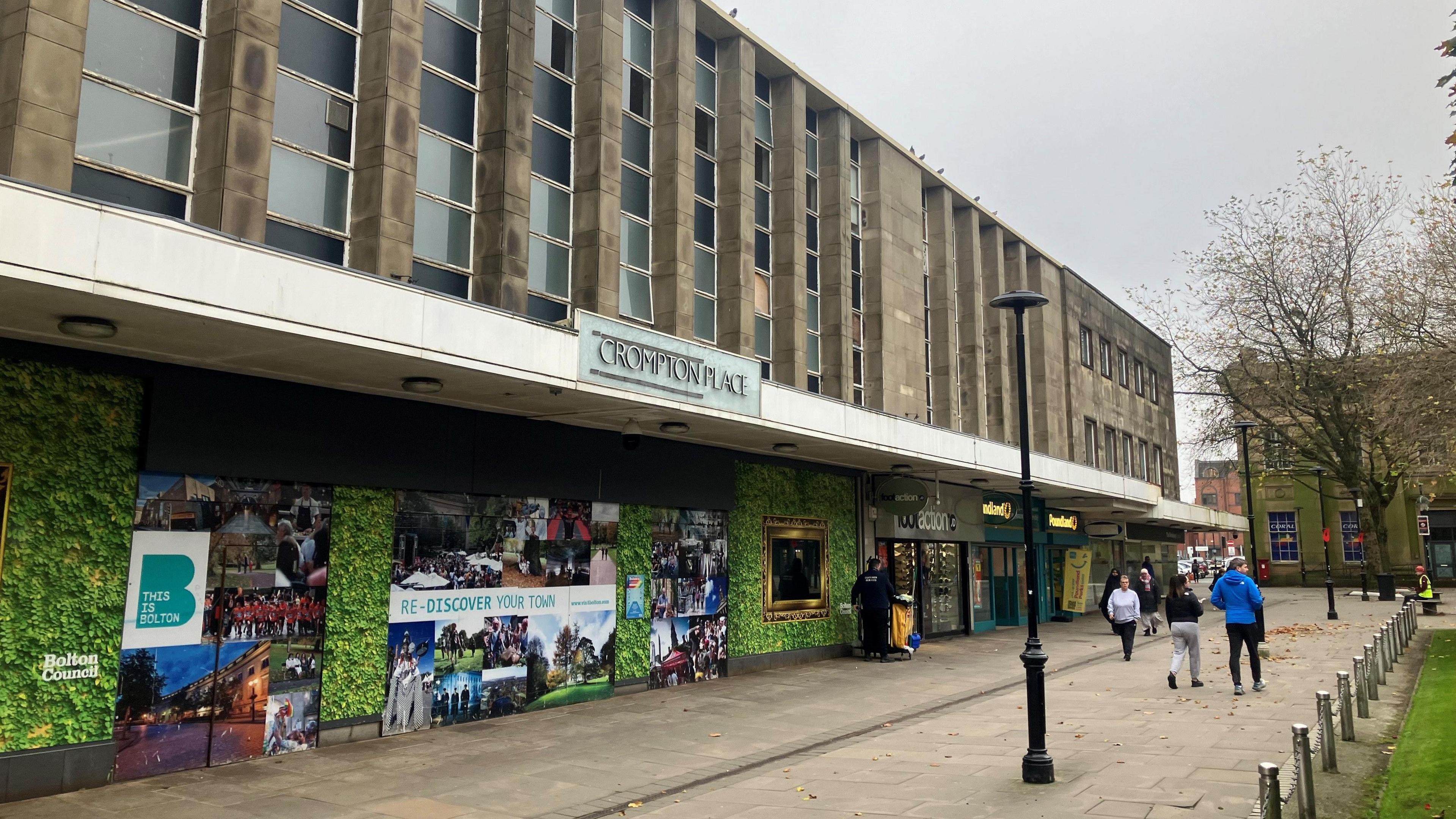 Crompton Place shopping centre. Bolton Council advert reads Rediscover your town. Shoppers walk past with metal posts on right hand side linked by chains running next to a grassy patch. There is a black lamppost in the middle of the path. 