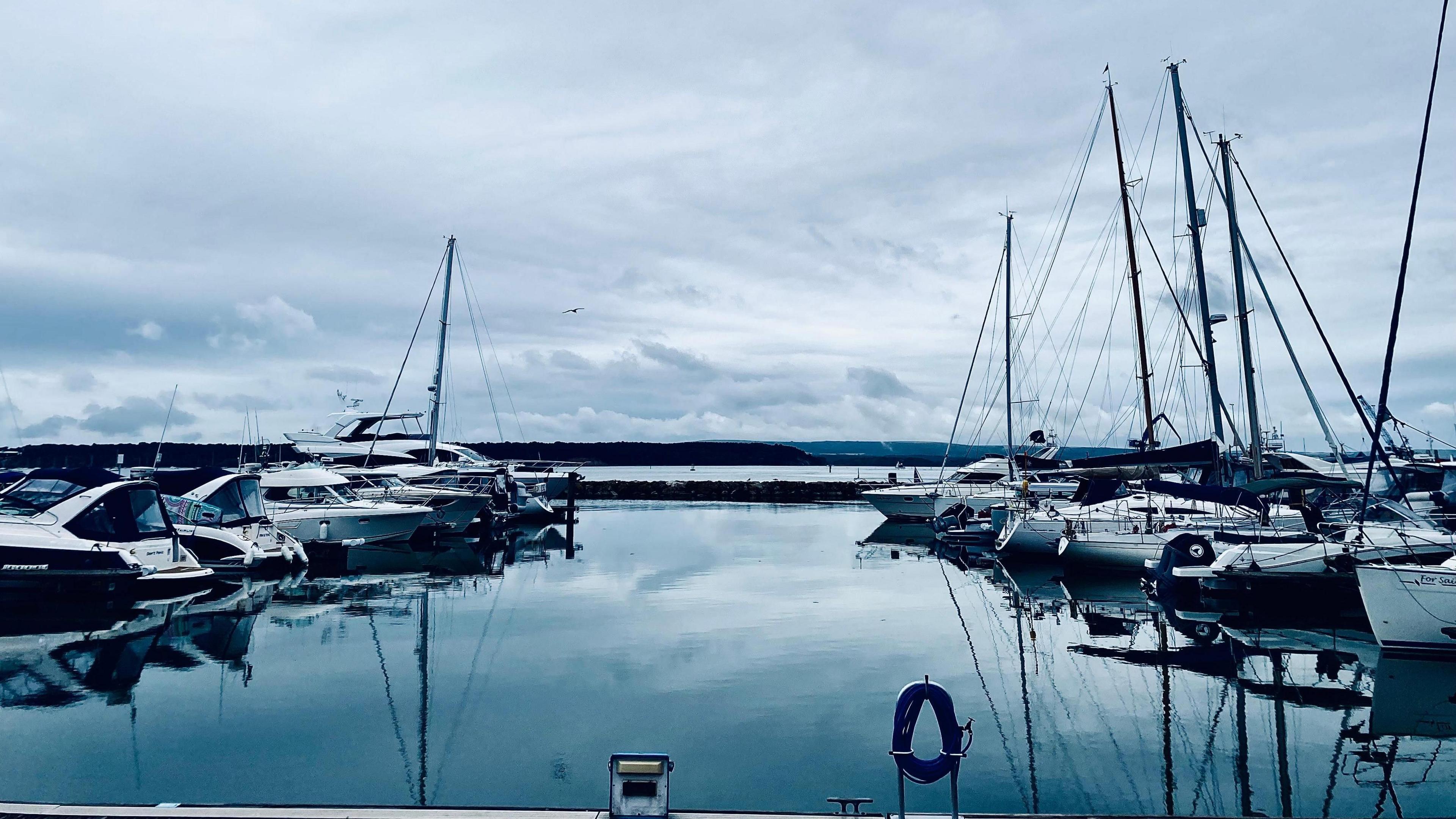 Two lines of moored yachts either side of the image sit on perfectly flat harbour waters which reflect the cloudy skies above. In the background breakwaters and the Dorset coastline 