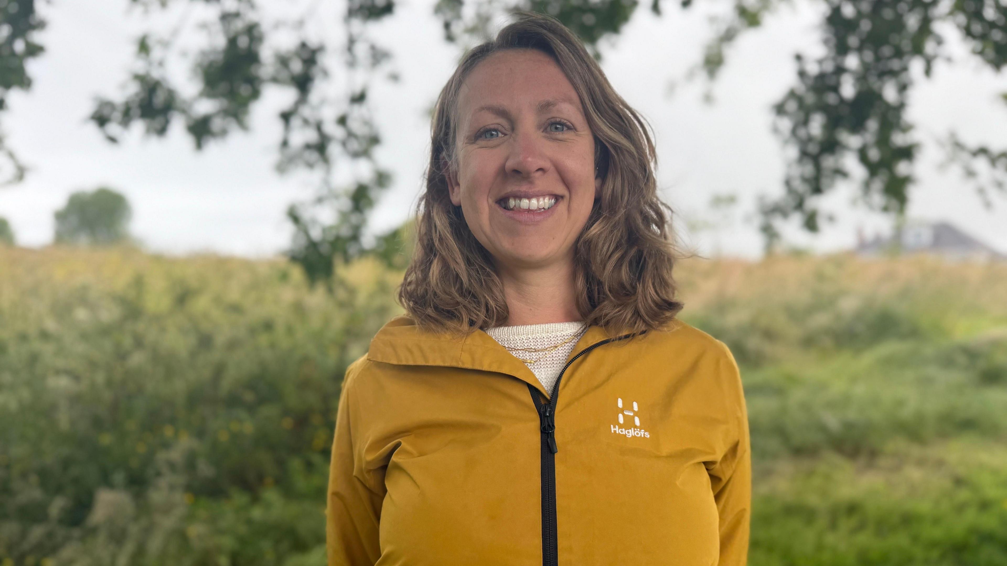 Julia Henny is standing under a tree and is smiling at the camera. He hands are held behind her back and she has shoulder-length curly dark blonde hair and blue eyes.