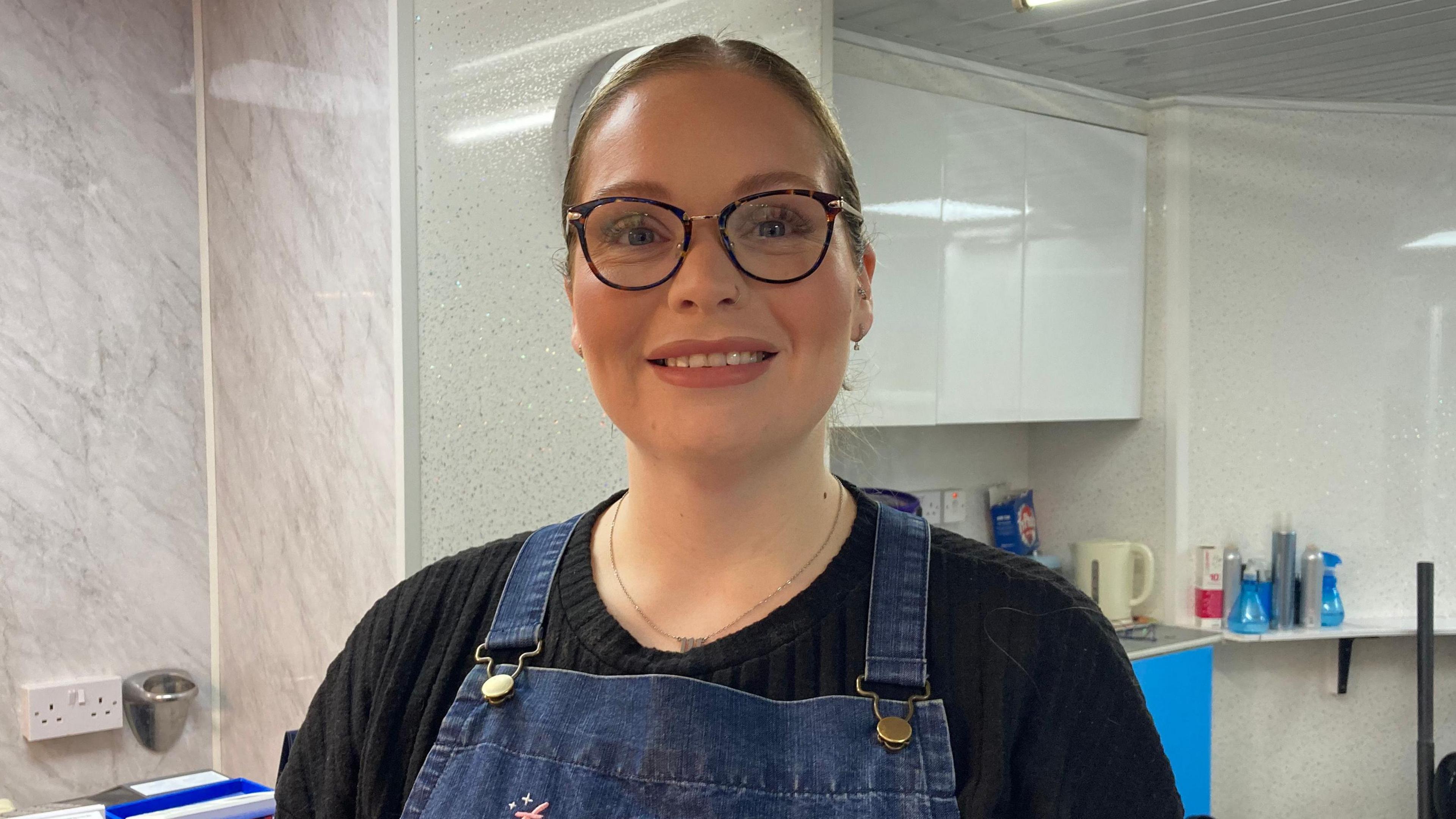 Victoria Hoskins is wearing a blue apron, a black shirt and glasses. She is standing in her hair salon with white cupboards behind her and equipment on worktops.