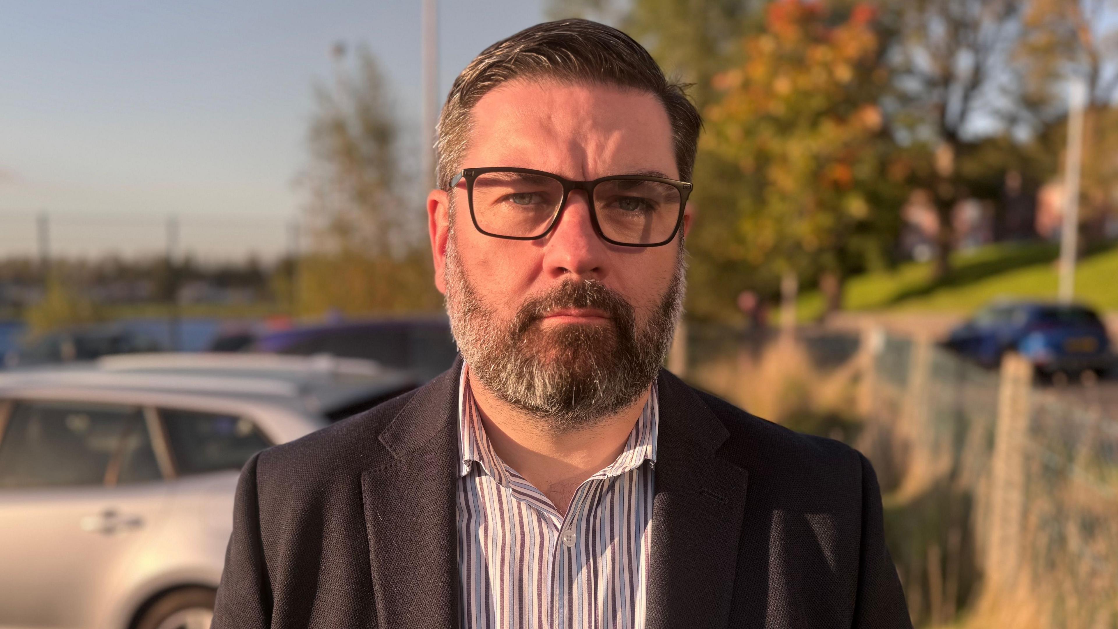 Niall McKenna has a neutral expression on his face as he looks at the camera. It is late evening, with the sun pointing towards his face. He has short black and grey hair, and is wearing a pair of dark framed glasses, a striped collared shirt and a black blazer jacket. He is standing in a car park, but the background is blurred.