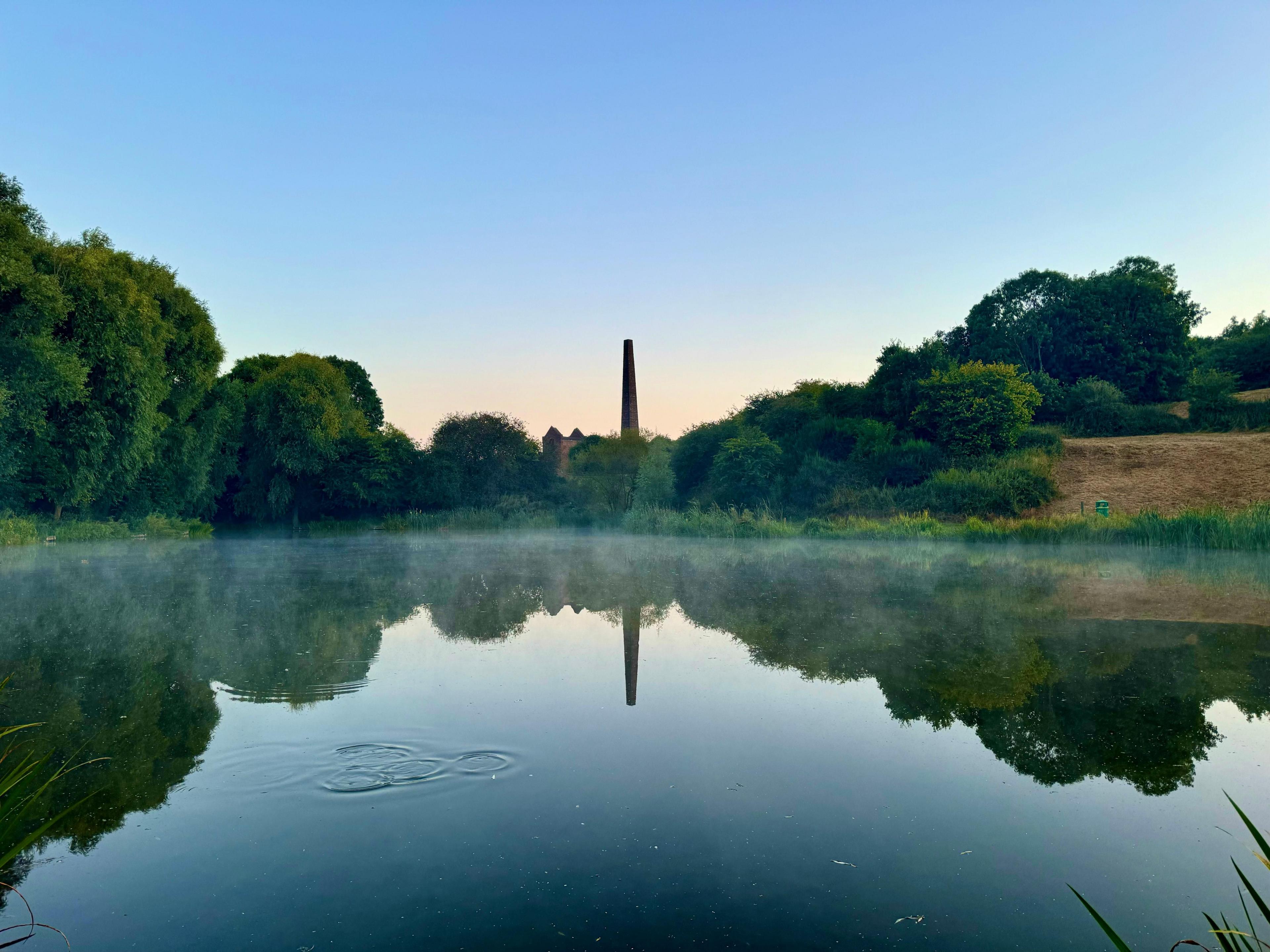 A Victorian-era brick chimney surrounded by trees is reflected in a still, slightly misty pond.