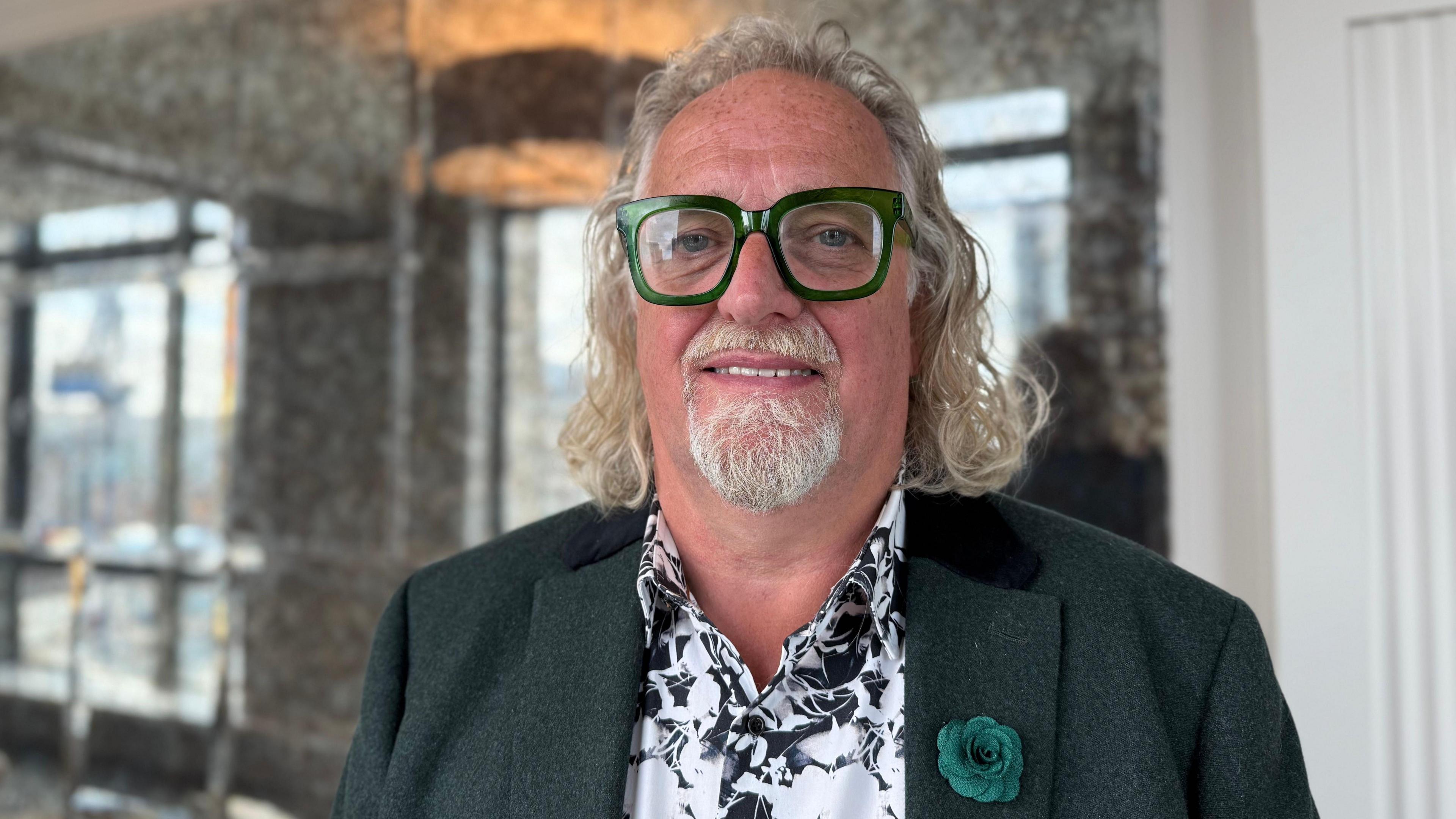 Belfast Night Czar Michael Stewart wears a green blazer over a white and green patterned shift. He wears large green glasses and has shoulder-length white hair.