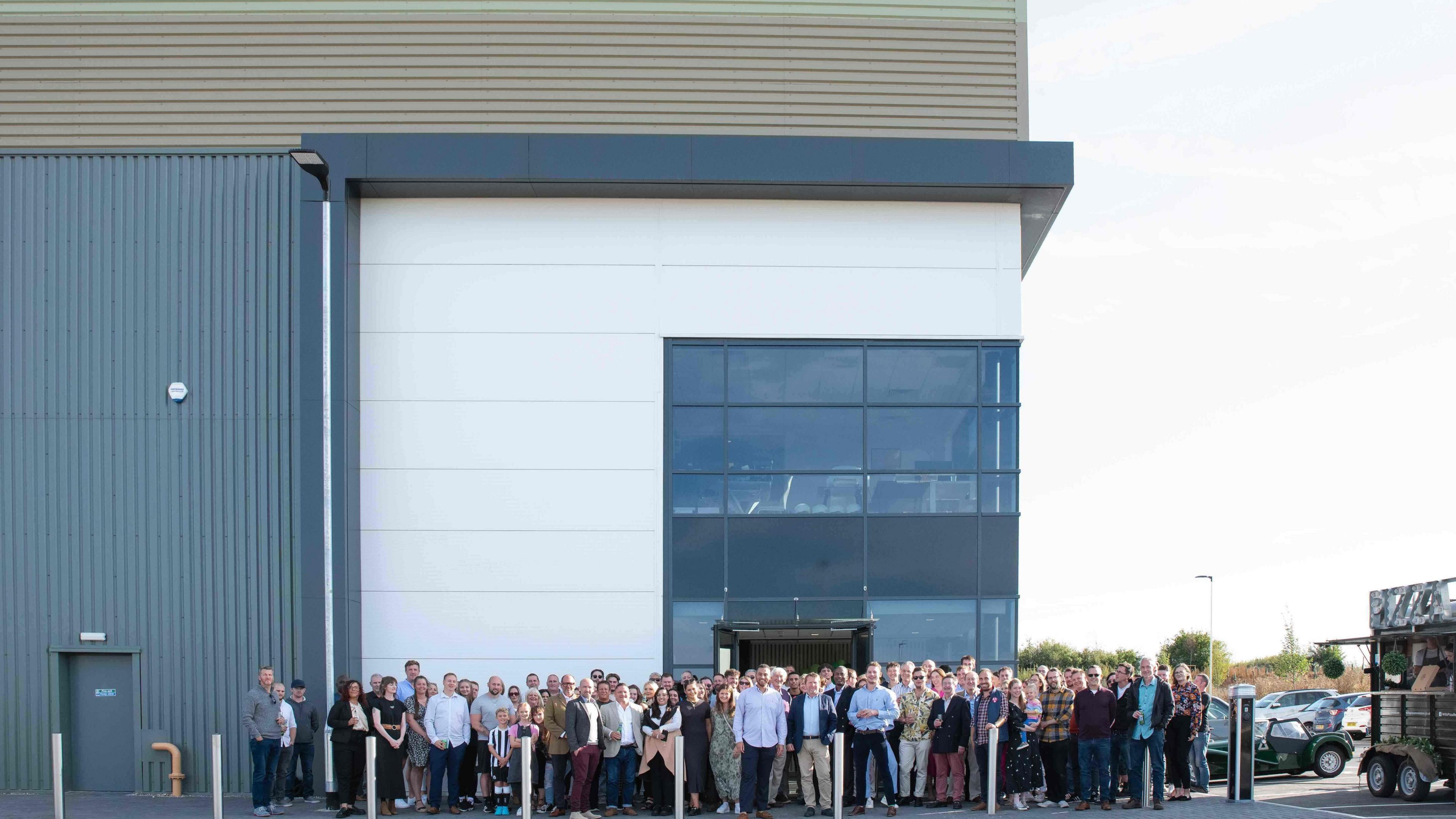 A crowd of people outside the entrance to a warehouse - a plain-looking box-like building. All are facing and posing for the camera.