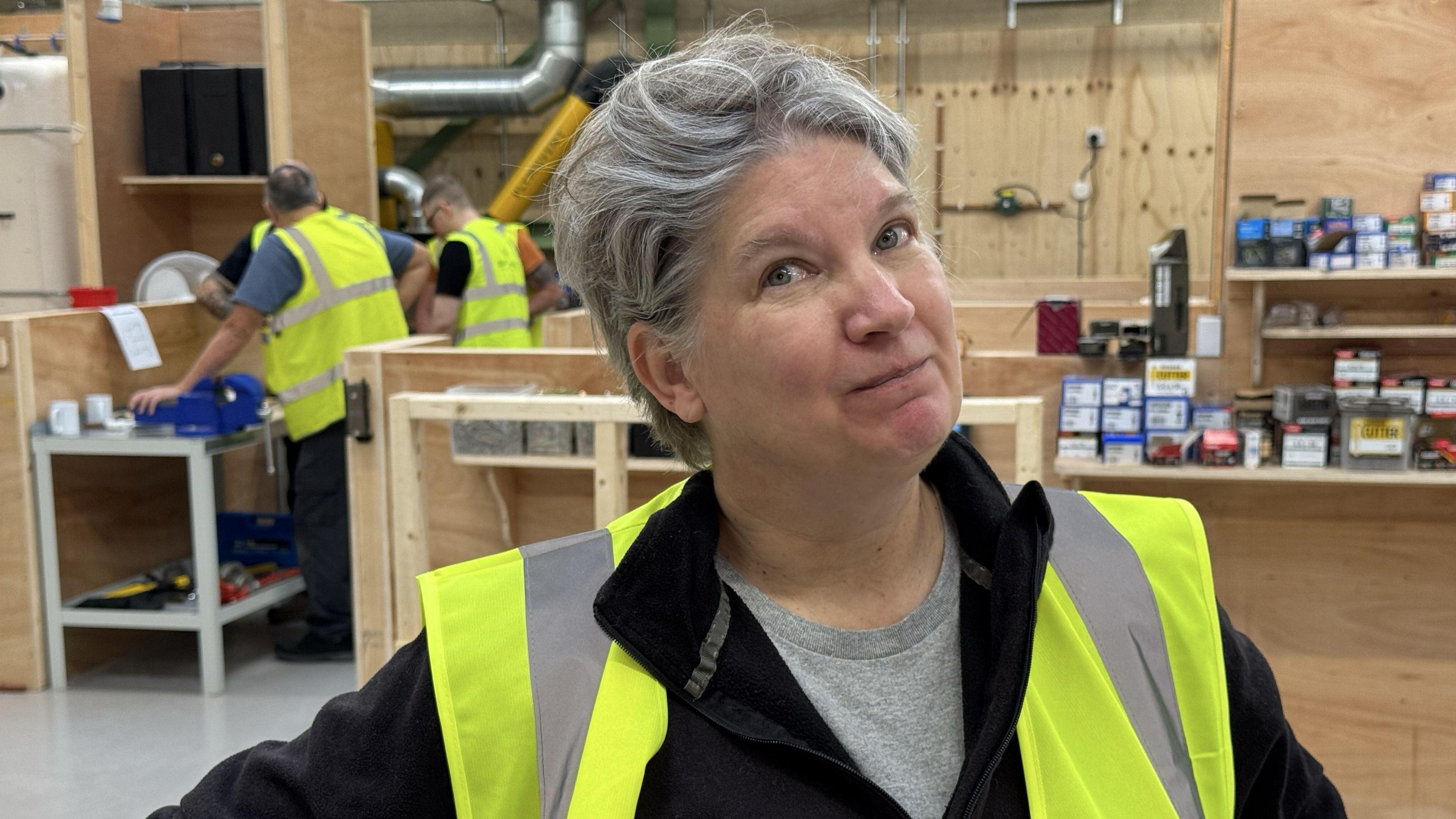 Marian Crenshaw-Austin, a woman wearing a black top and high visibility jacket, is standing in what appears to be a woodwork studio. There are three other people in the background who are also wearnig high visibility jackets. Shelving in the background appears to contain boxes of DIY or construction materials.