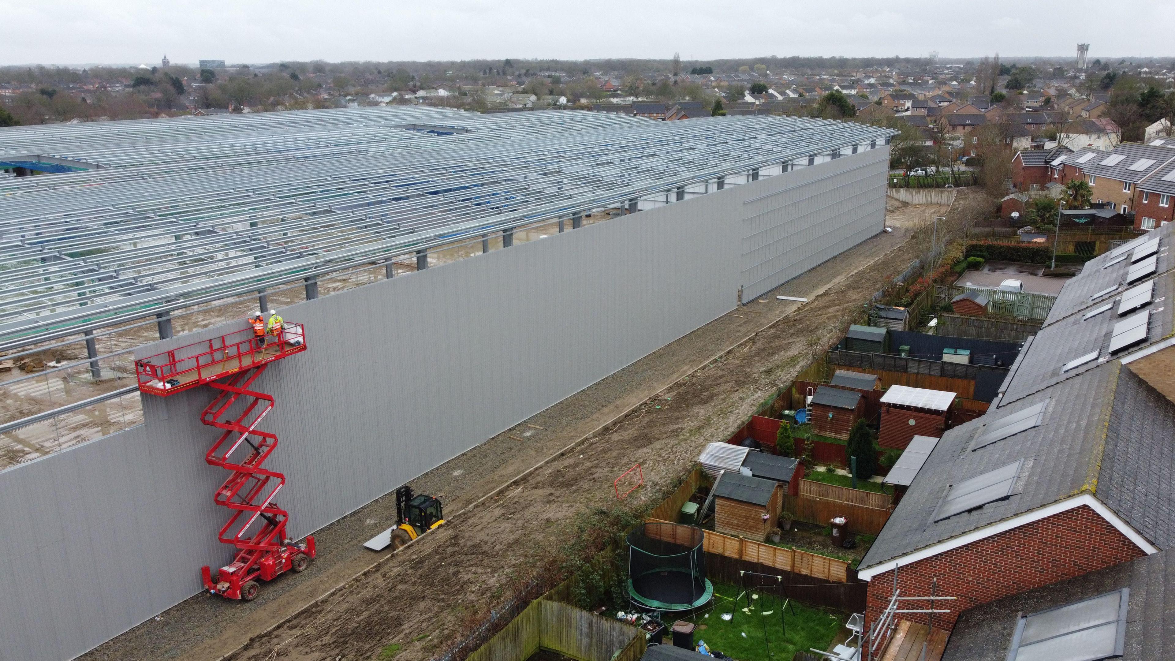 A drone shot of the incomplete warehouse - a grey structure that stretches across the frame several metres in front of a housing estate. The estate backs directly onto the warehouse land.