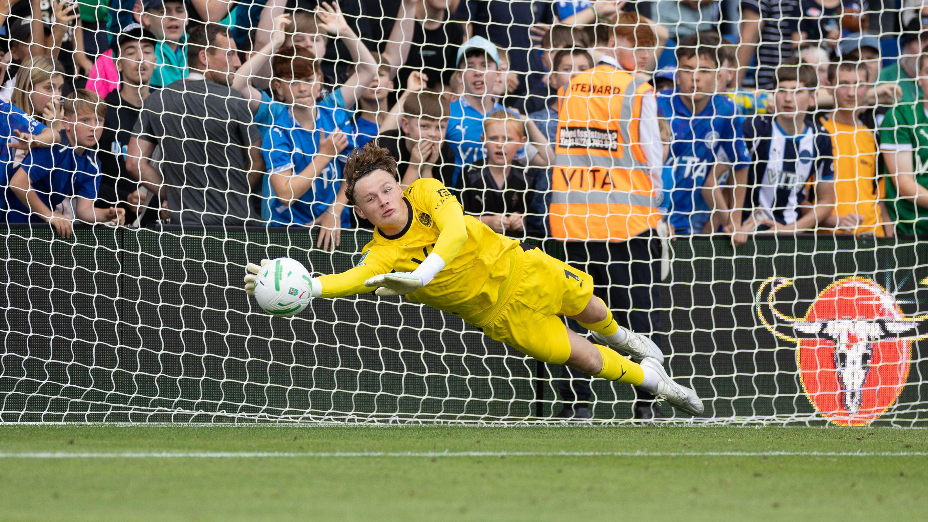 Oli Whatmuff dives to save the ball in his yellow Stockport County goalkeeping kit during their game against Doncaster Rovers in the EFL cup