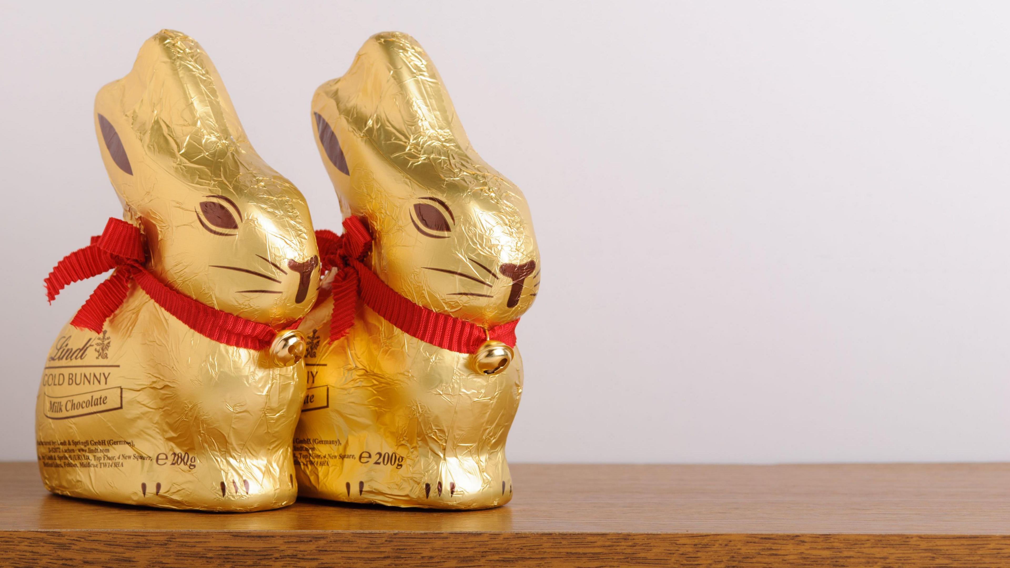 Two Lindt Easter chocolate rabbits next to each other on a wooden table with a blank beige wall behind. The rabbits are wrapped in gold coloured foil and have a red ribbon around their necks with a bell on.