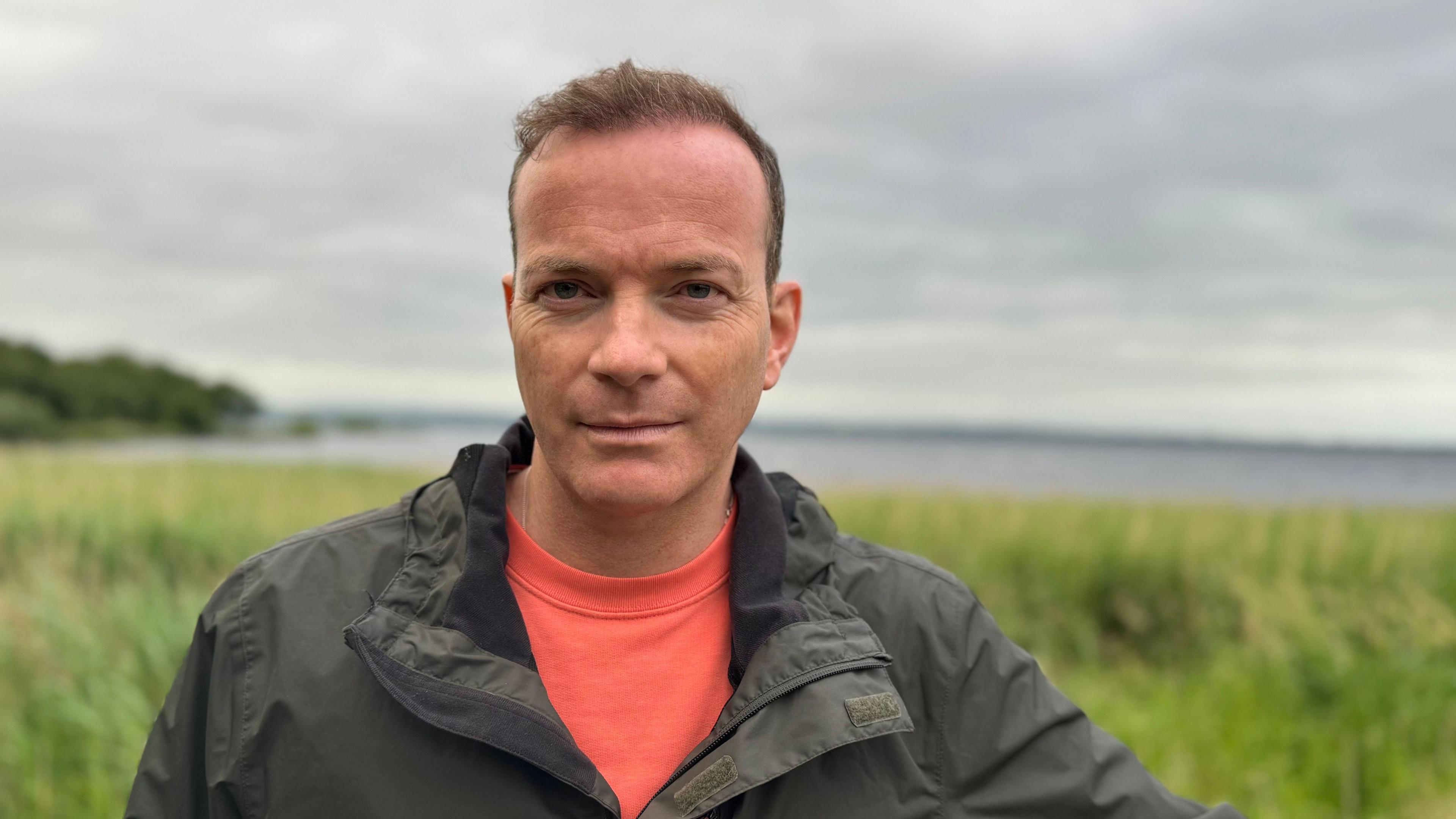 Simon Chambers. He has short brown hair, wearing a khaki raincoat and orange t-shirt. He is standing on the shores of Lough Neagh.