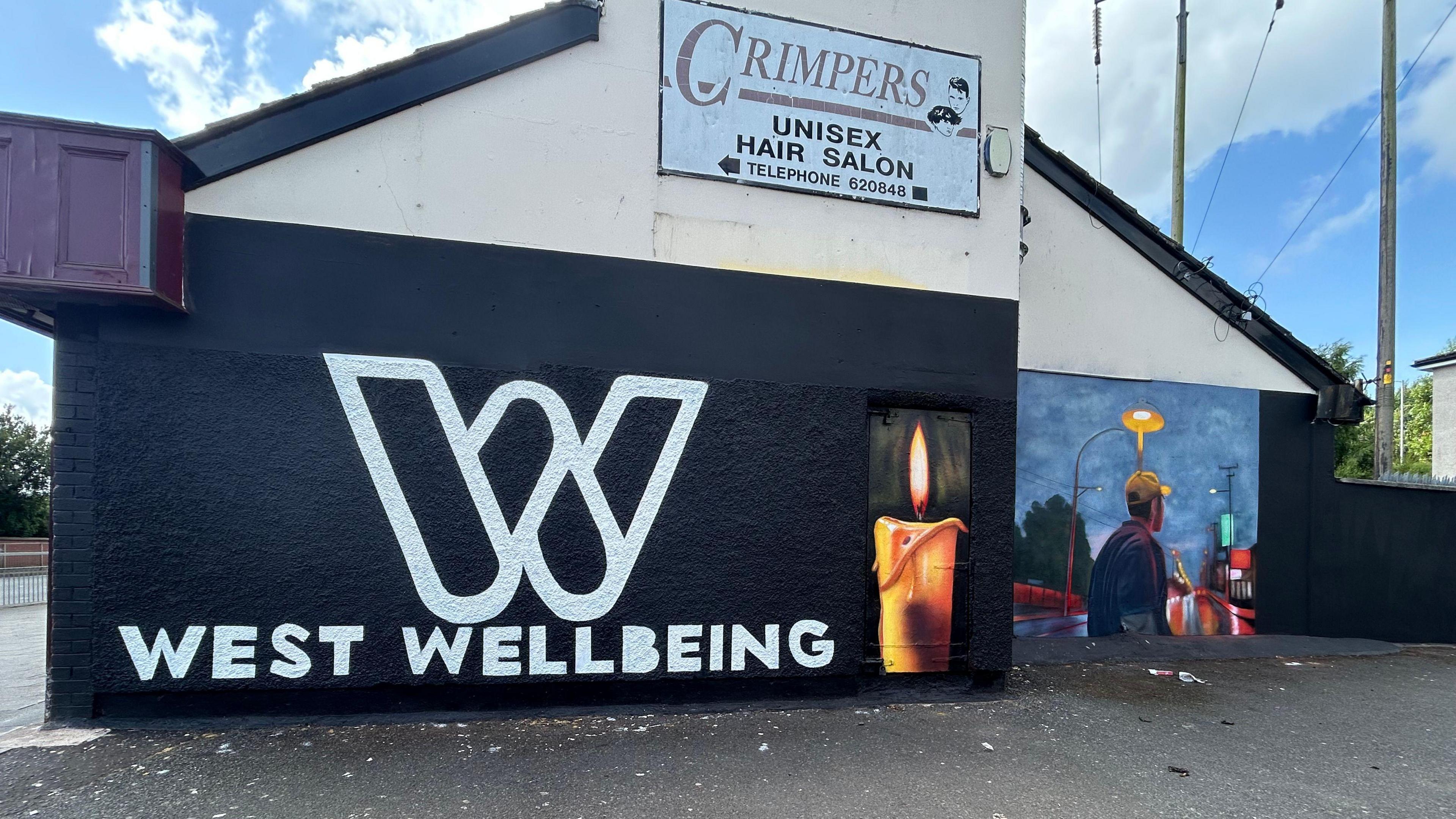 The side of a gable wall is painted black with white lettering stating, West Wellbeing. A painting of a young man is on the next wall.