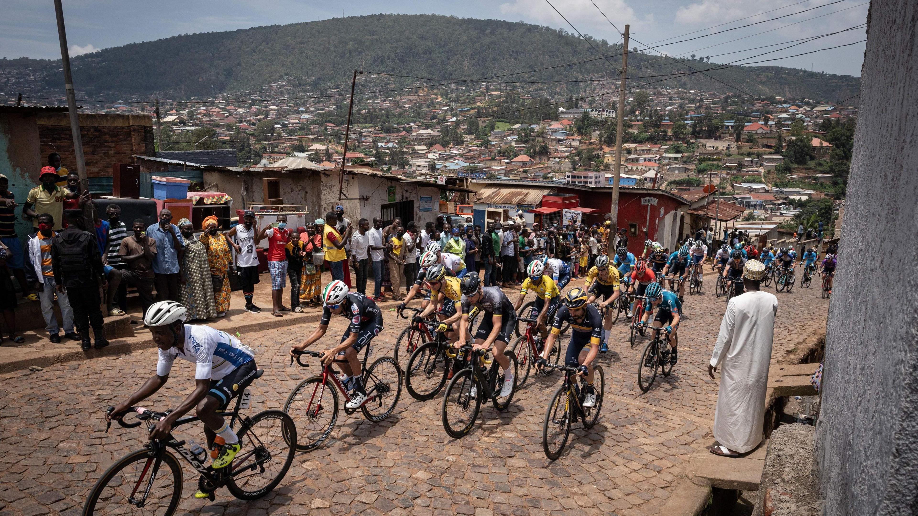 Cyclists competing at the Tour du Rwanda in 2022 travel up a hill through an urban area as people watch on, with more buildings rising up on the side of a hill in the background.