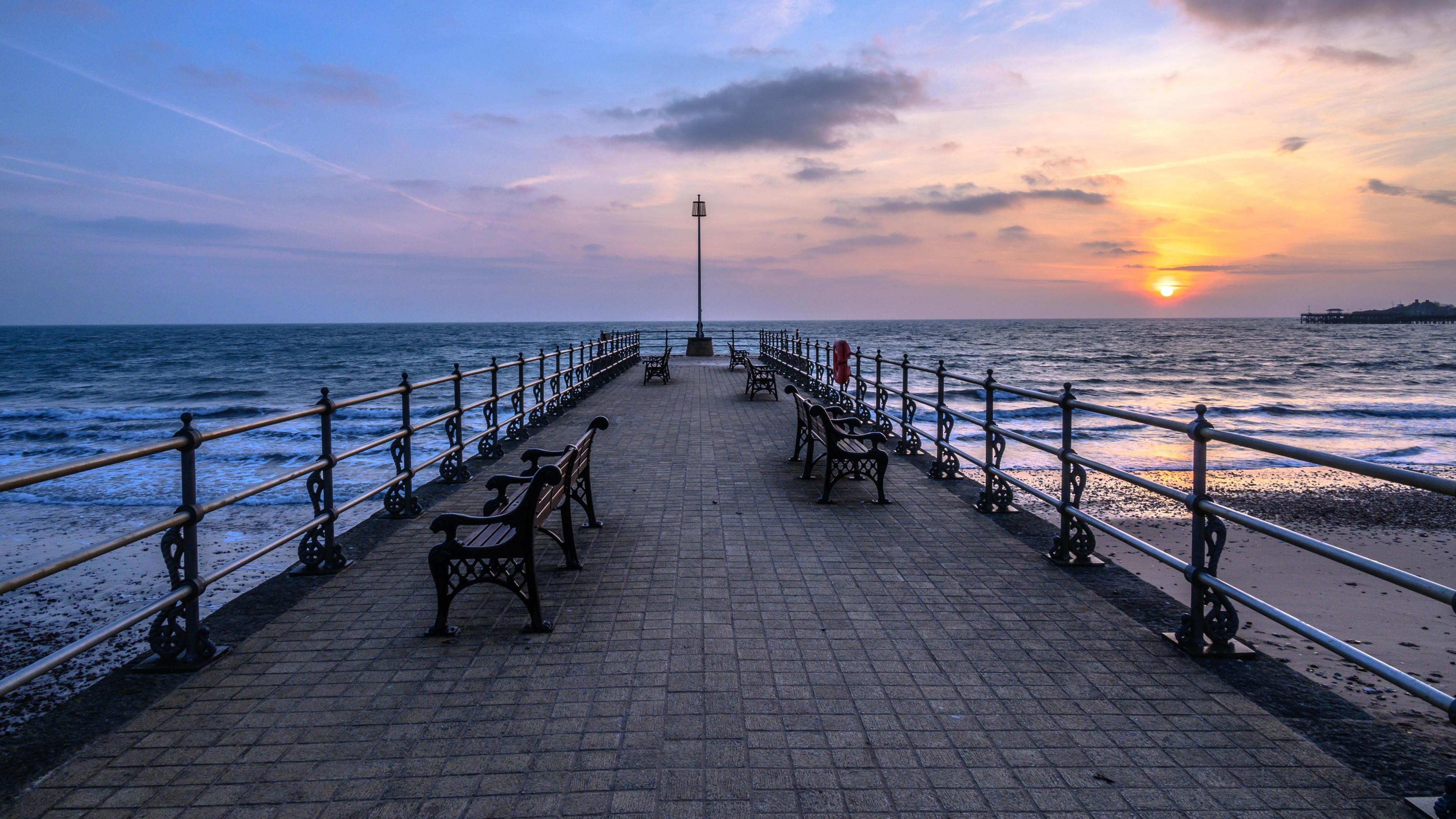 Looking directly down the pier, which has a circular end. There are benches on each side and a life float fastened to the railings. The paving is small square tiles. The sun is setting in the distance.