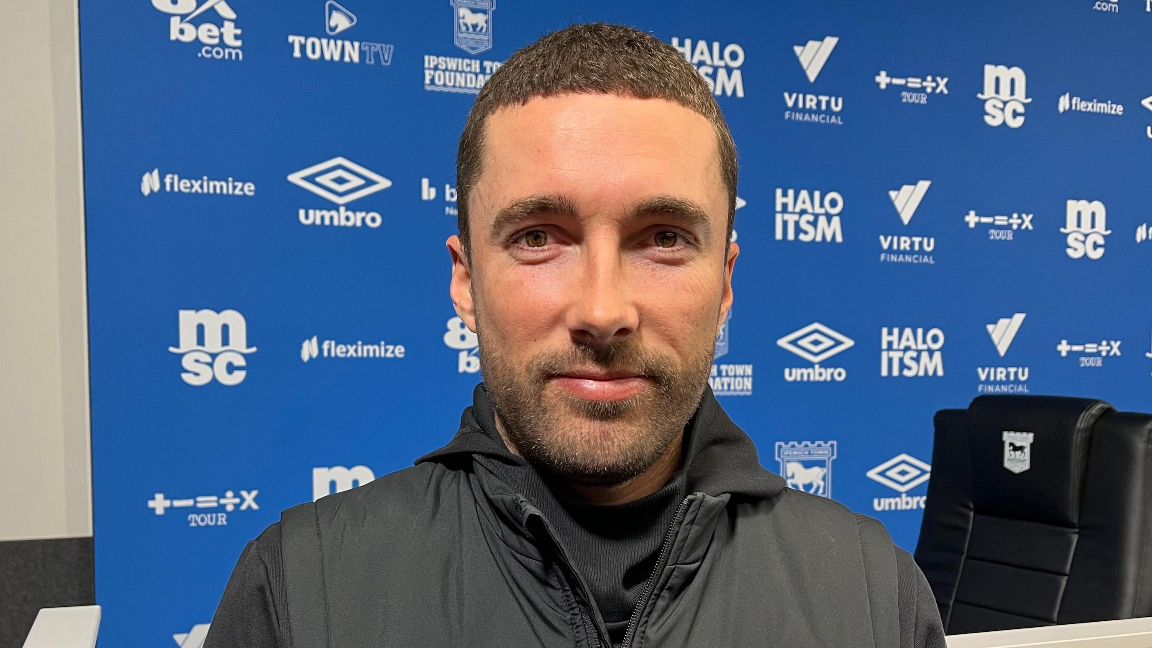 Joe Sheehan, the team's manager. He is looking straight at the camera. He has short black hair and a short black beard. He is wearing a black gilet and top. He is smiling. He is standing in front of a blue hoarding which is enblazoned with the names of sponsors.