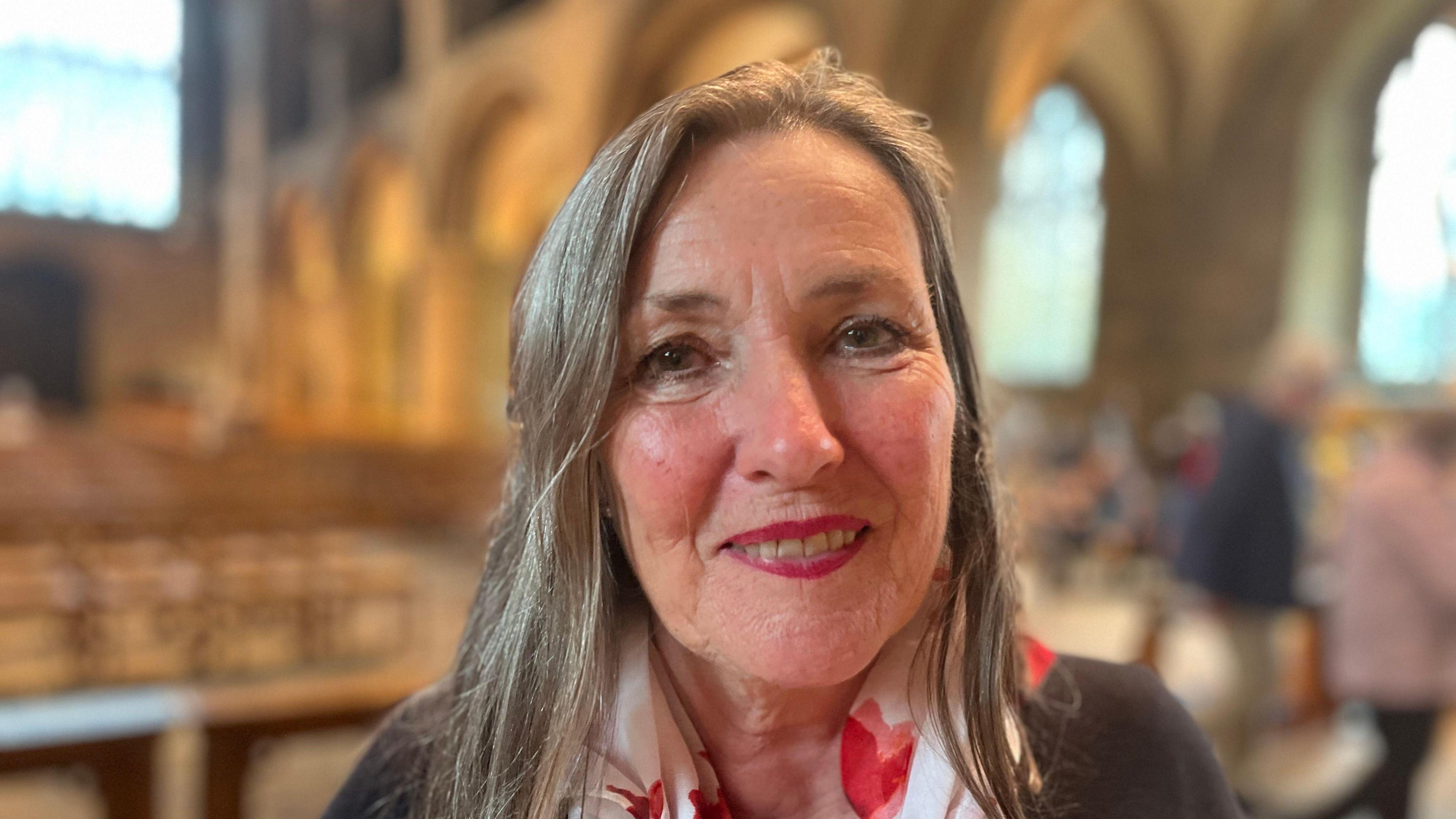 Tansy Forster. She has long grey hair, and is wearing a black jumper with a red and white scarf. In the background, out of focus, is a church.