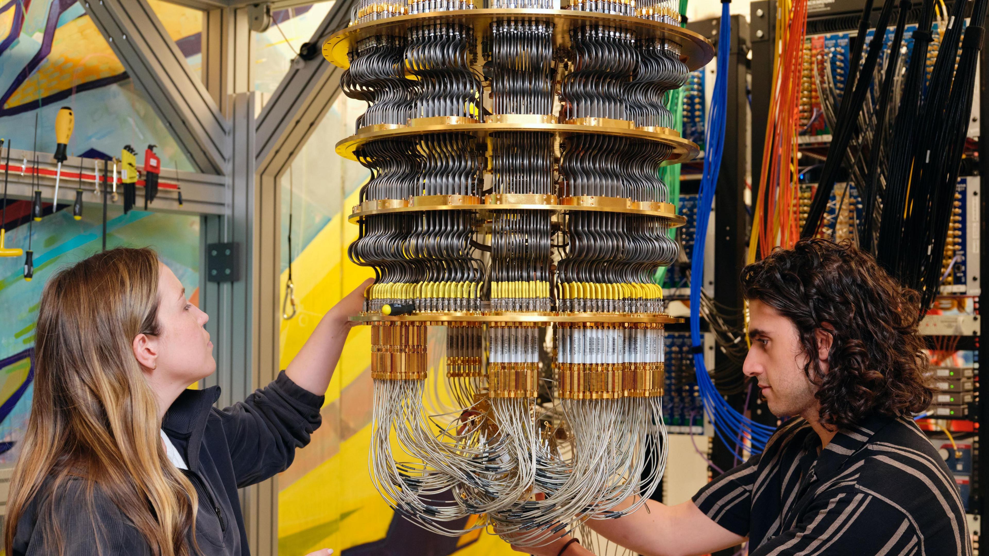 Google staff, a woman to the left and a man to the right work on the cryostat which holds the chip and keeps it very cold. The cryostat losley resembles  a chandelier  made of cascading thin metal tubes.