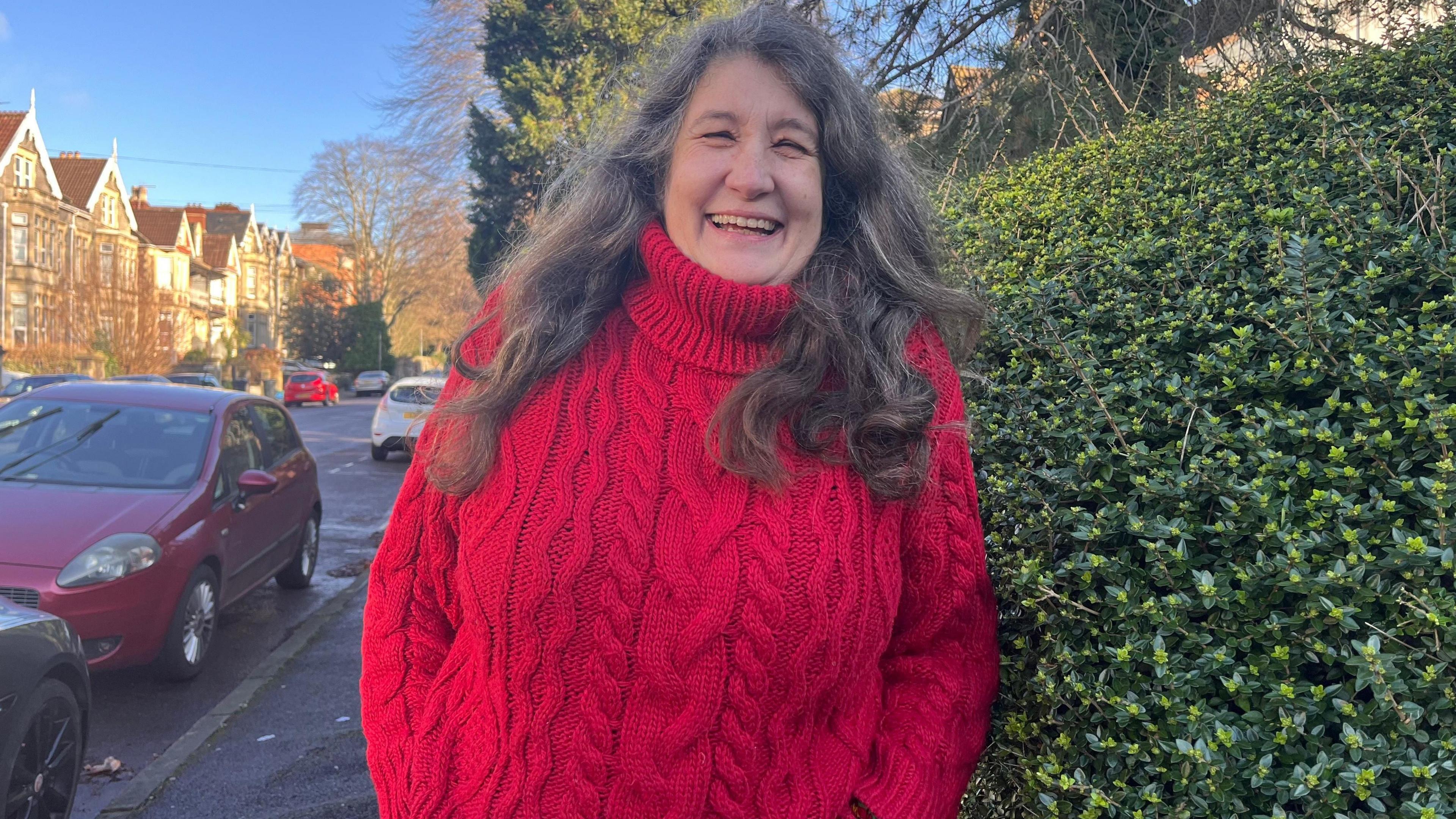 Kitty Wilson smiling, she has long, curly dark hair and is wearing a red knitted jumper. She is pictured outside, standing on the pavement next to a hedge and a row of parked cars.