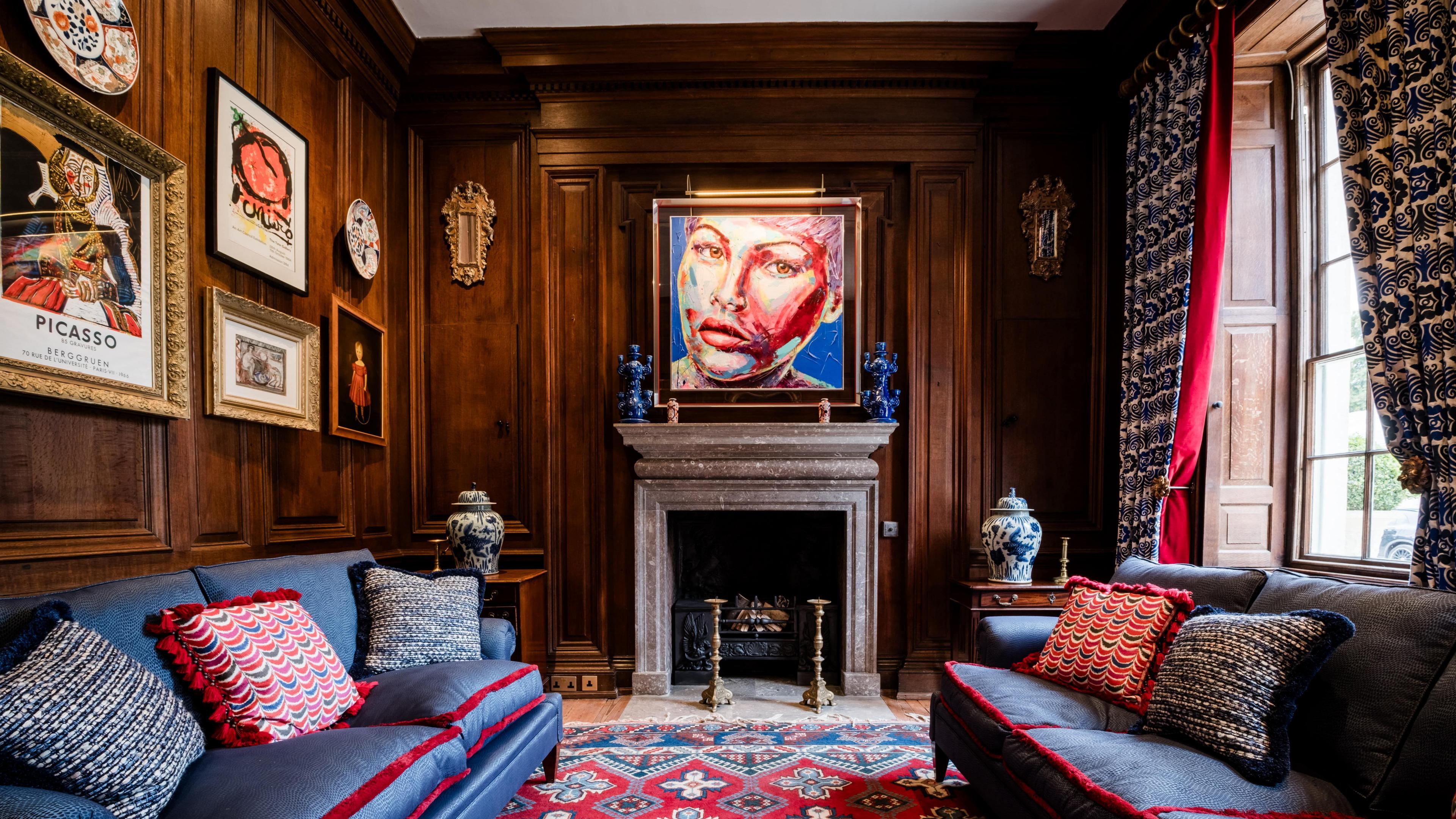 The "Oak Room" at Hotham Hall. The entire room is covered in dark wood panelling. A traditional fire place is in the centre of the image with a colourful painting of a female face hanging above it. There are two blue sofas with red trim in the room and both have colourful cushions on top of them.