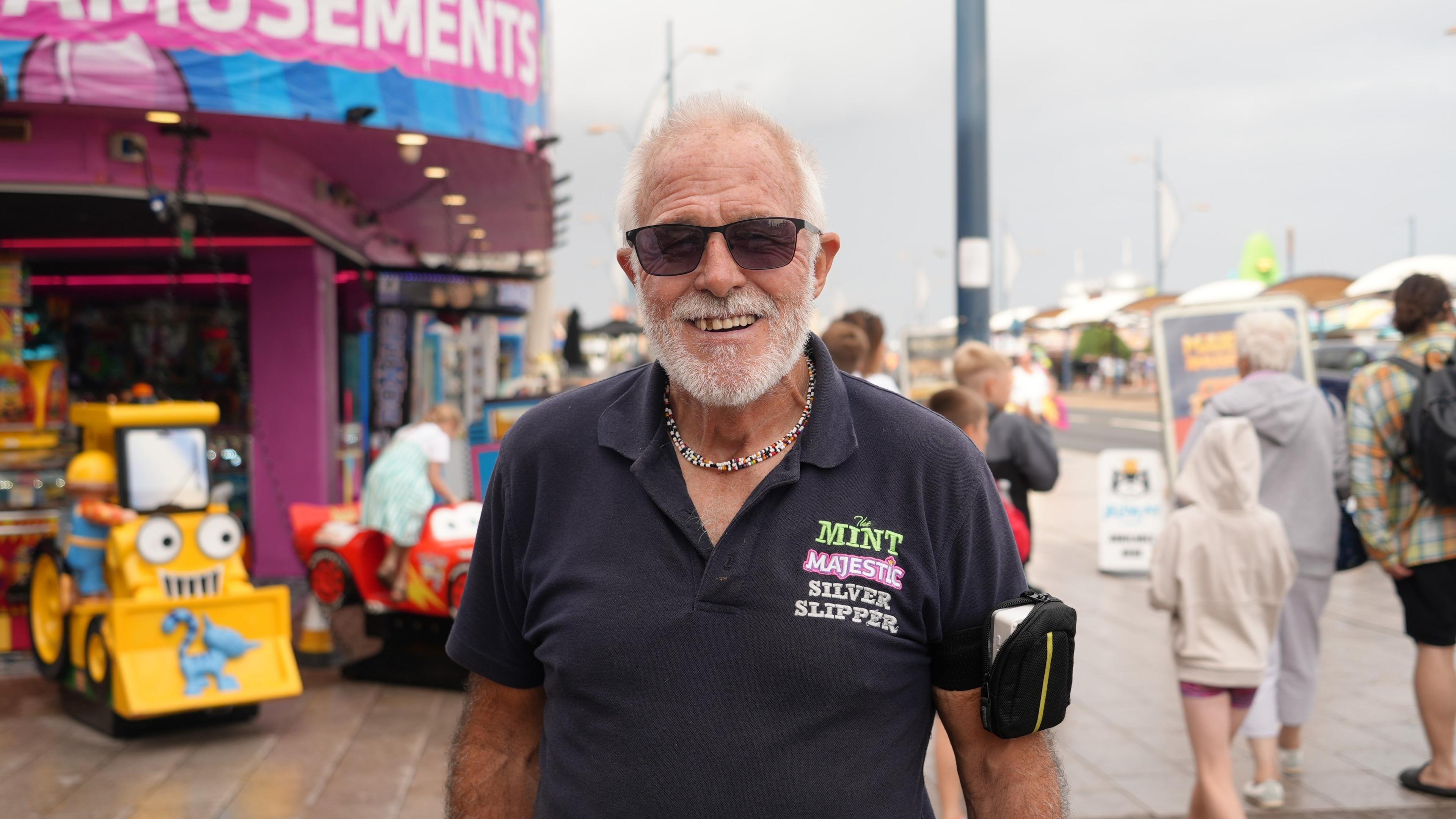 John Stowers is smiling to the camera - he has a grey white beard and wears dark glasses and a dark navy polo shirt
