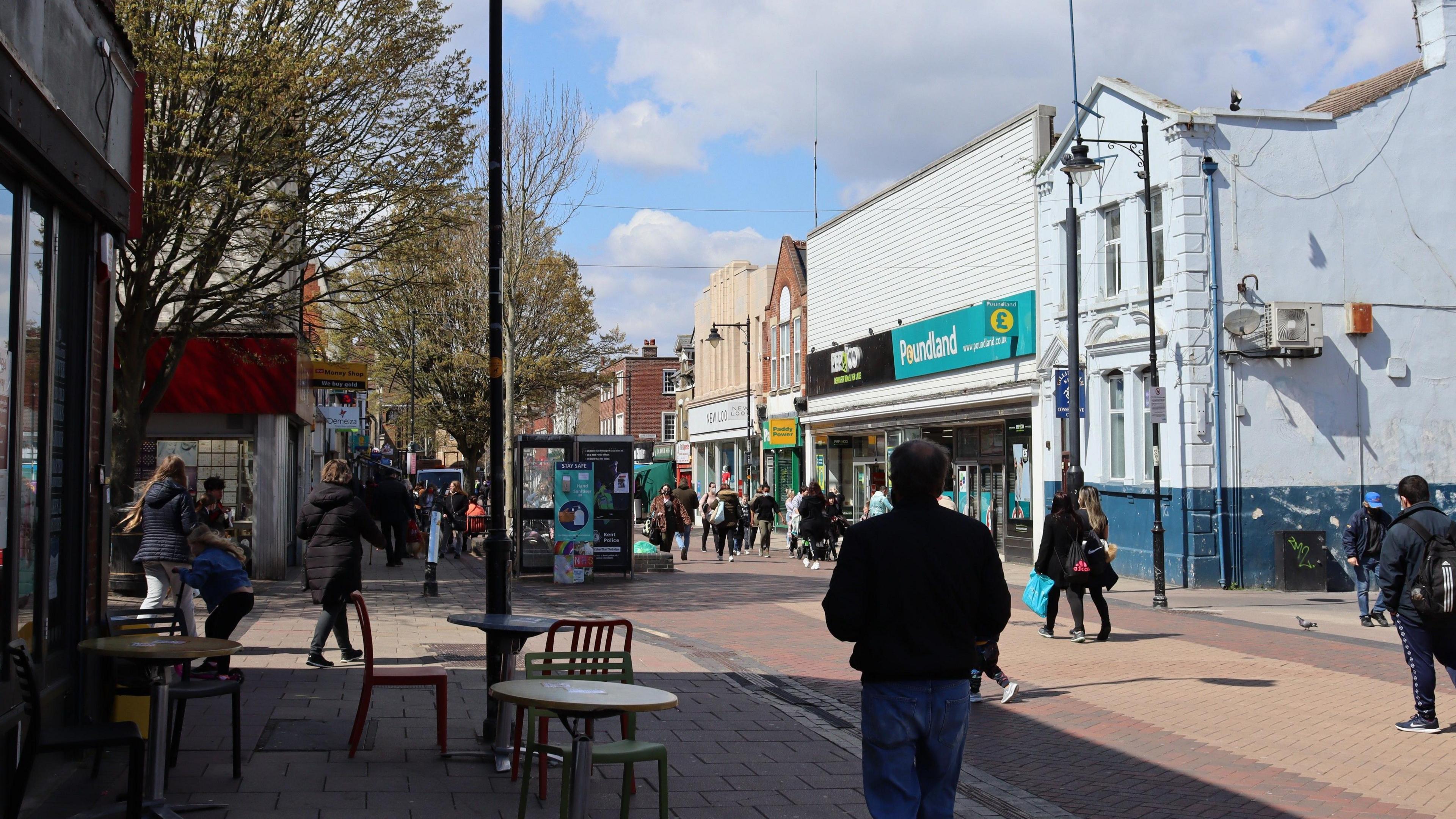 There are tables and chairs on the pavement and shoppers walking by on a sunny day in Gillingham