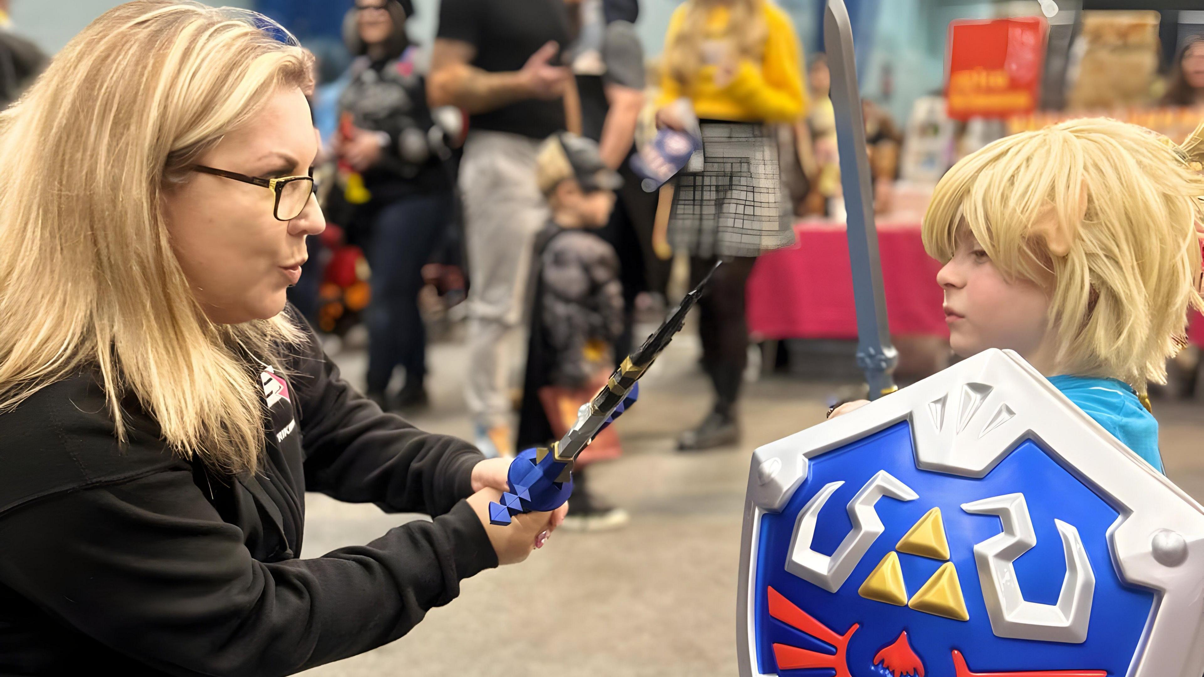 Zoe Crofts has blonde hair and is holding a toy sword. A child dressed as Link from Zelda is stood in front with a shield and sword. They are at a convention, a child is dressed as Batman behind them.