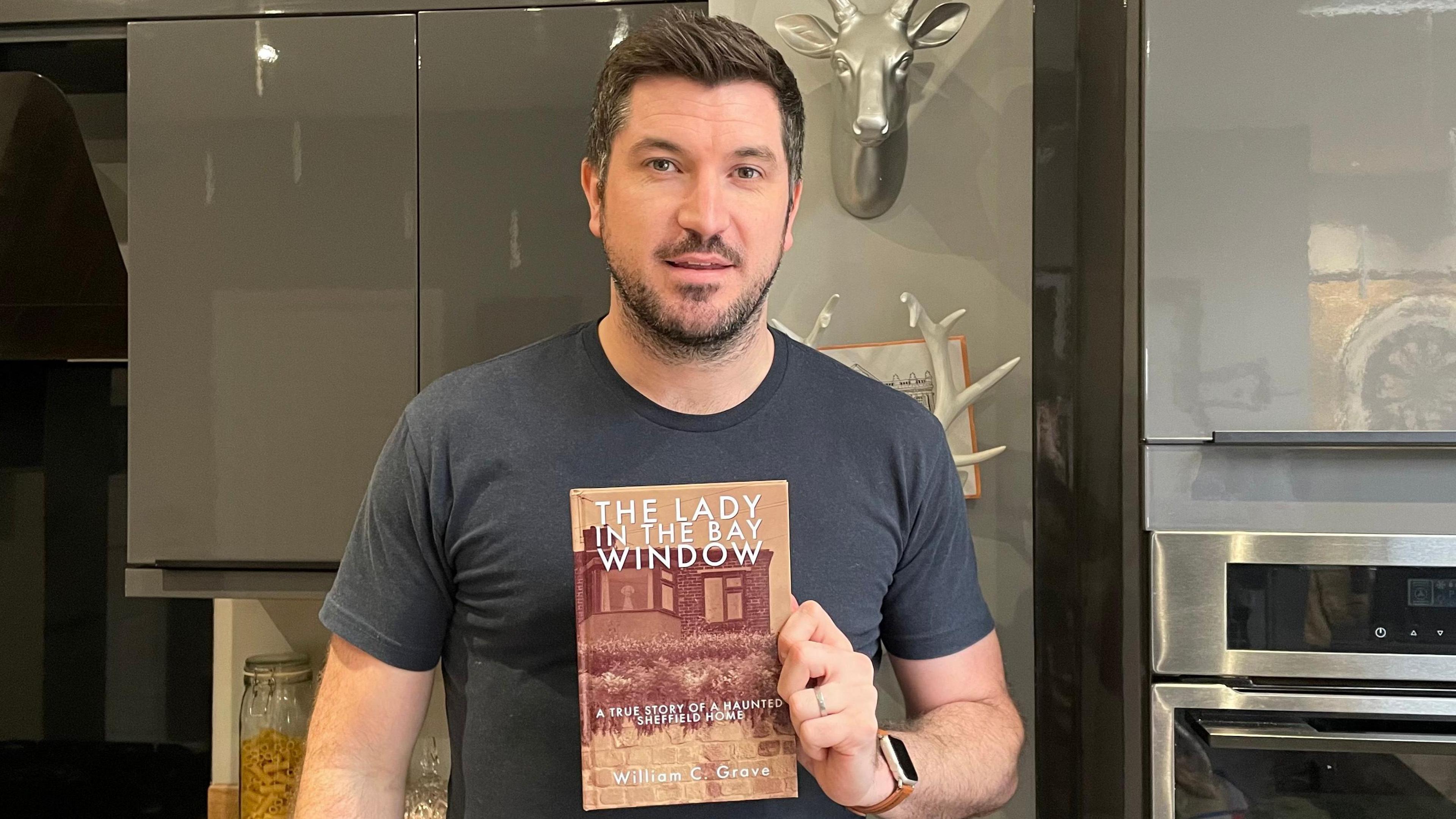 Liam Kippax is stood holding a copy of the book 'The Lady in the Bay Window.' On the cover is a photo of a house with the ghost of an elderly woman in its window.