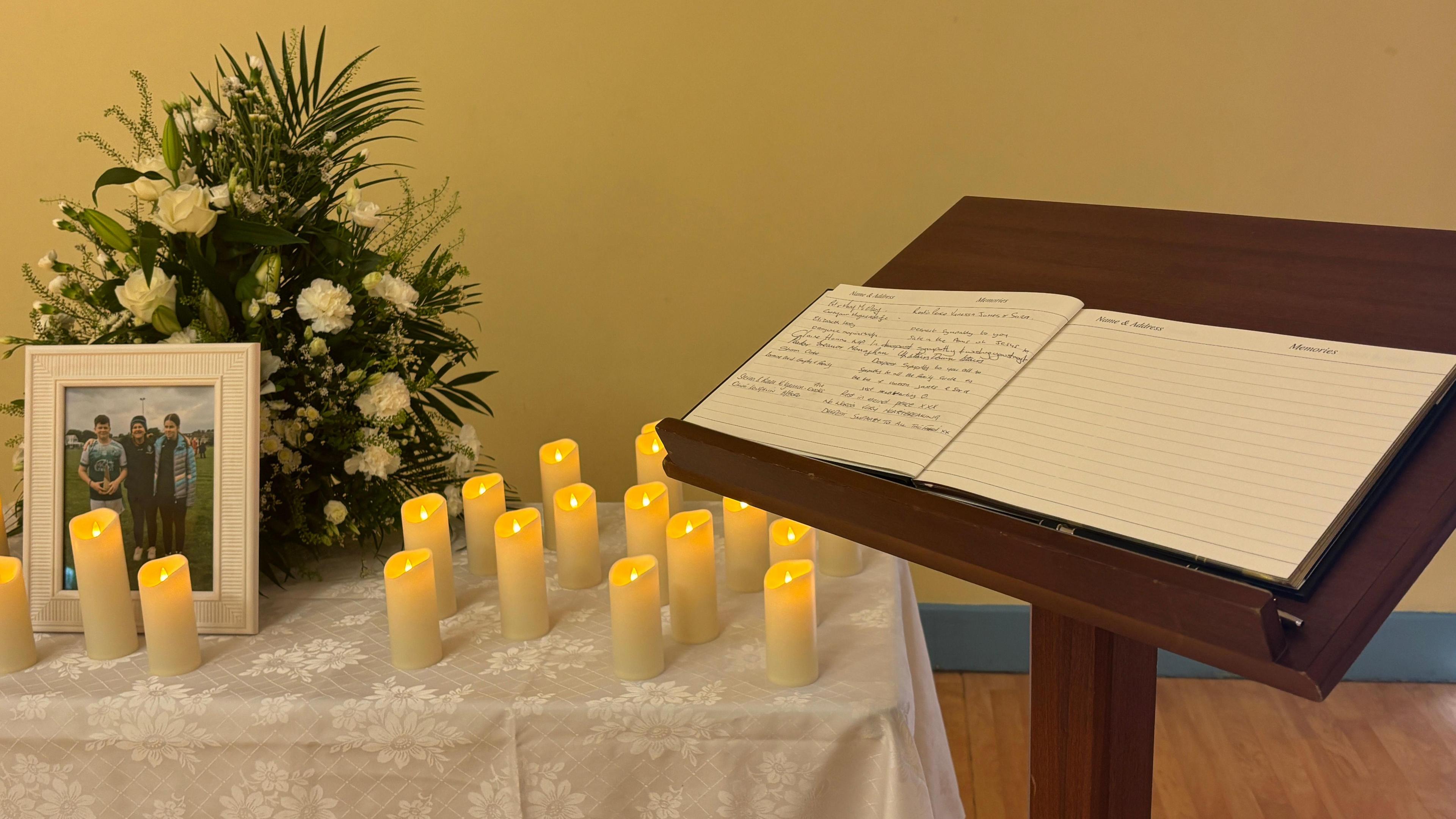 A book with handwritten messages is on a lectern. Next to it is a table upon which are a floral display, more than a dozen electrical candles and a photograph of Vanessa Whyte with her children Sara and James.