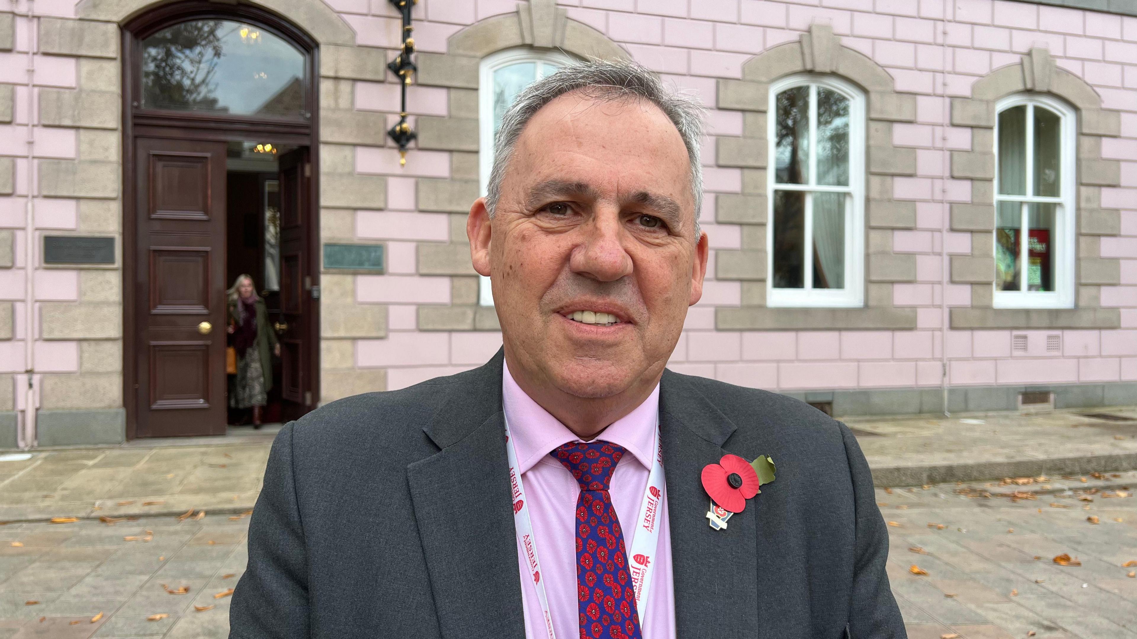Constable Andy Jehan, a man with short grey hair. He is wearing a dark grey suit, a pink shirt and a navy blue tie with lots of little red poppies on it. He has a white lanyard around his neck with red writing on it, which says "Government of Jersey"'. There is a poppy on his lapel. He is standing in front of Jersey's States Assembly building. To the left of the image, someone is walking through the door to the chamber.
