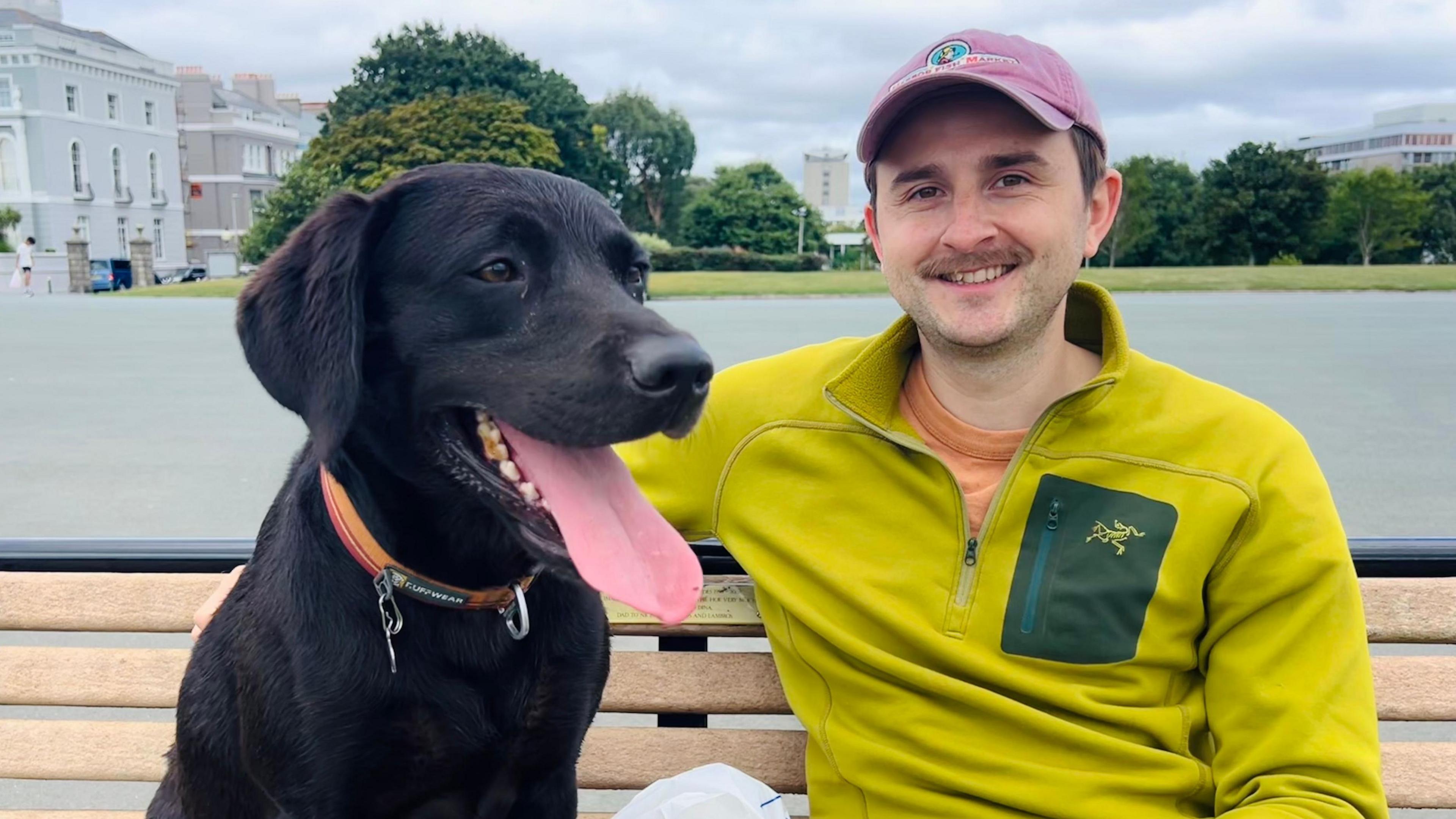 Black labrador Logie sits on a bench next to his owner James Westgate. James is wearing a lime green fleece, a pink baseball hat and is smiling at the camera. Logie has his tongue hanging out.