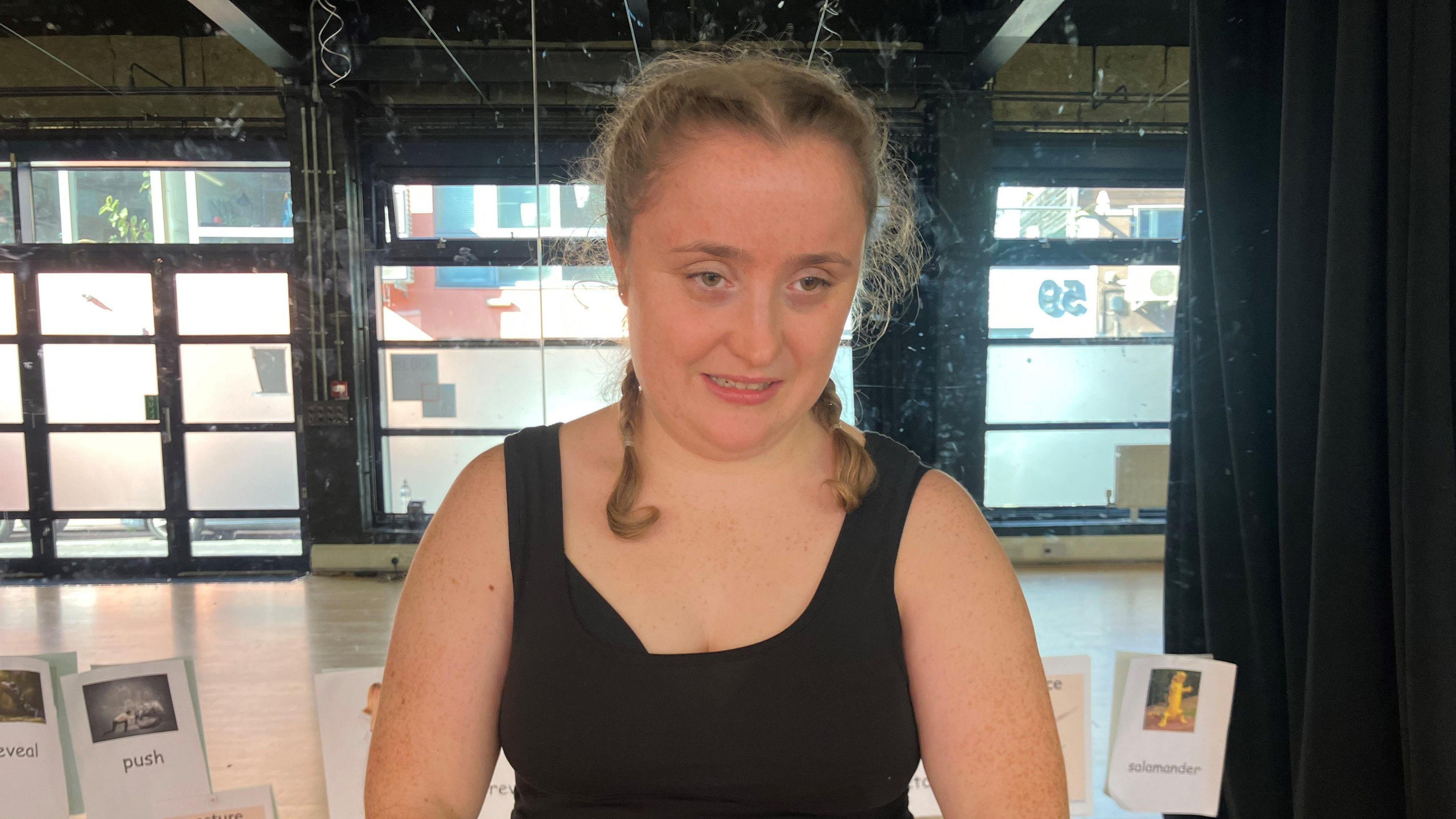 A woman wearing a black sleeveless t-shirt. She is stood in front of a mirrored wall with pictures attached to it.