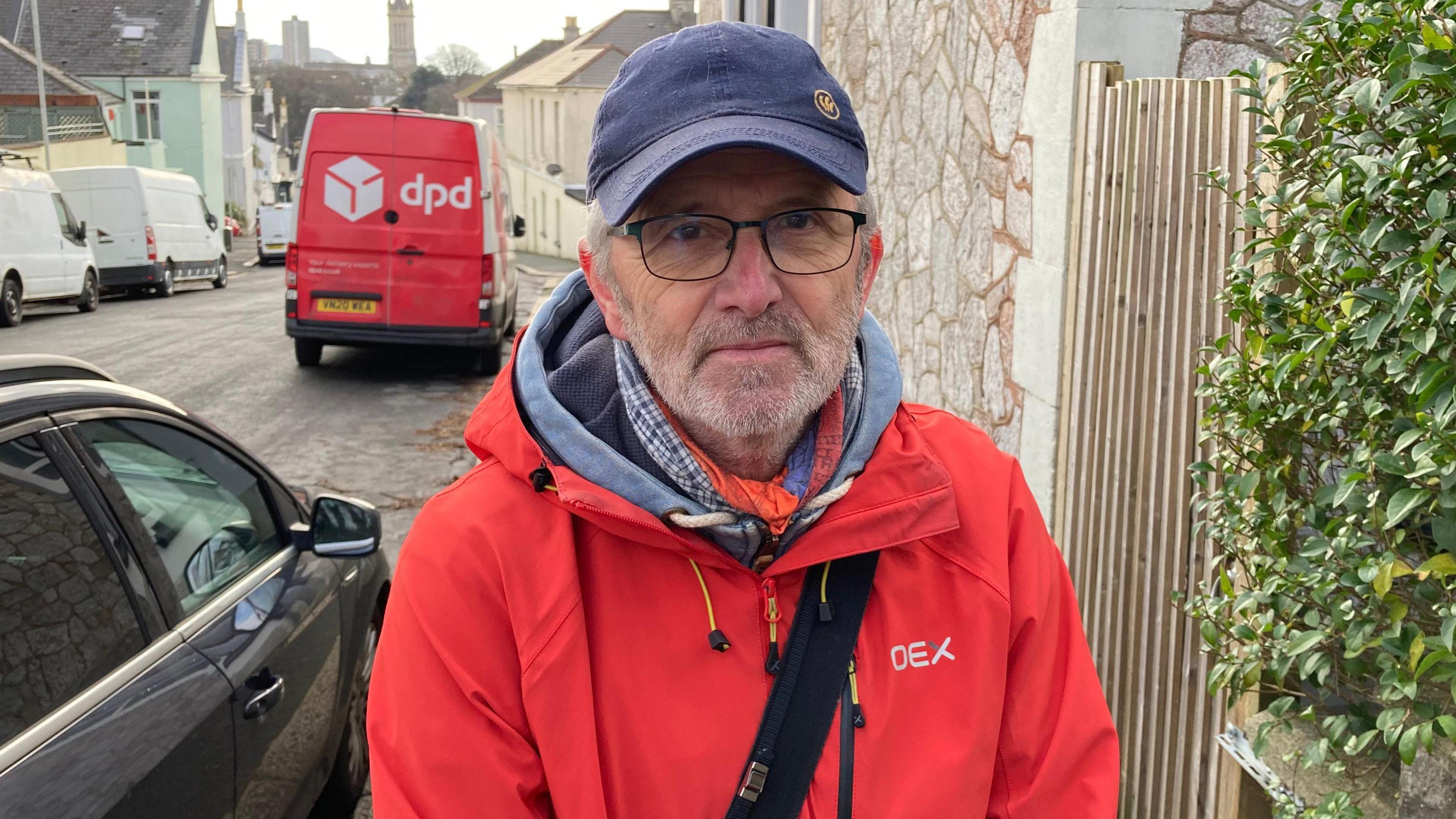 Simon Alston wearing a red coat and with a black backpack strap across his chest. He has a navy blue baseball cap on and is standing on a pavement next to a garden's hedge.