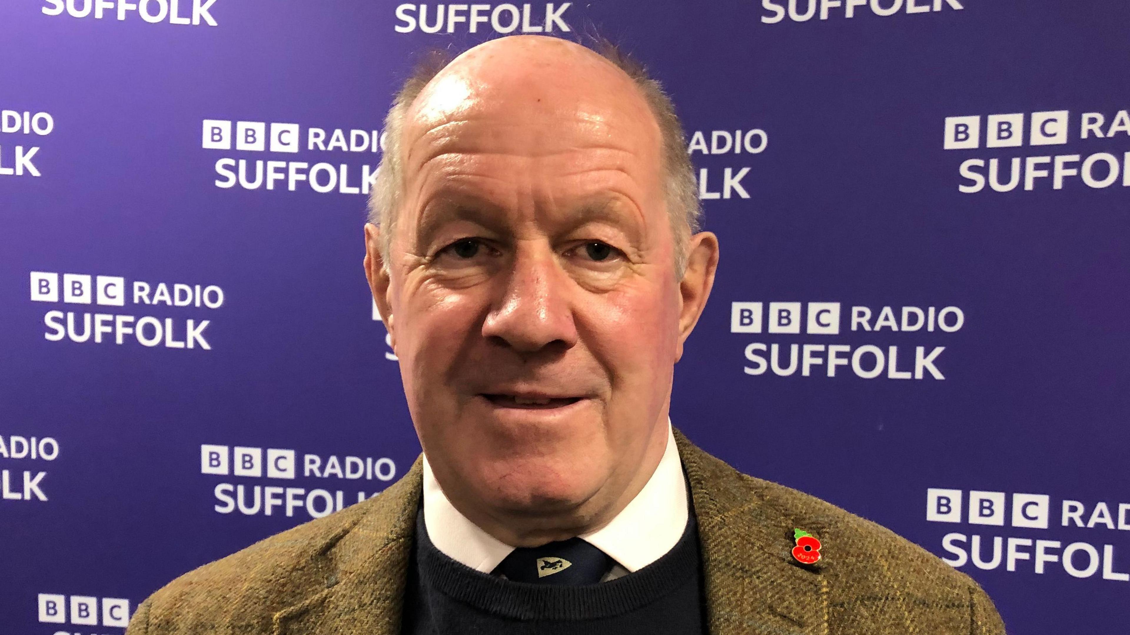 Tim Passmore smiles at the camera while standing in front of a purple coloured wall with the BBC Radio Suffolk logo written on it. He is largely bald with some grey hair and he wears a tweed jacket with a black jumper and white shirt and tie underneath.