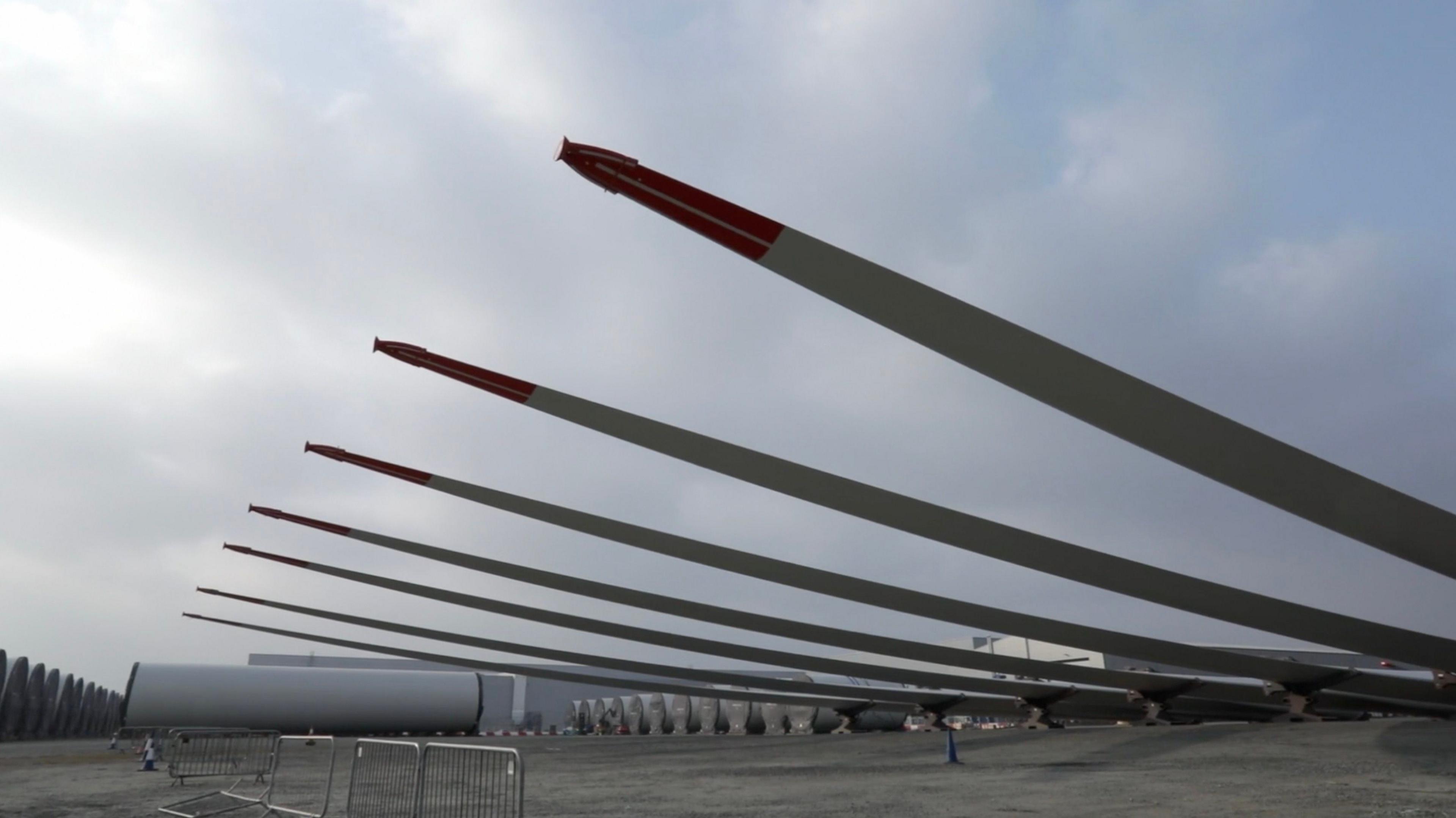 Seven large turbine blades are propped-up at a large open factory site. 