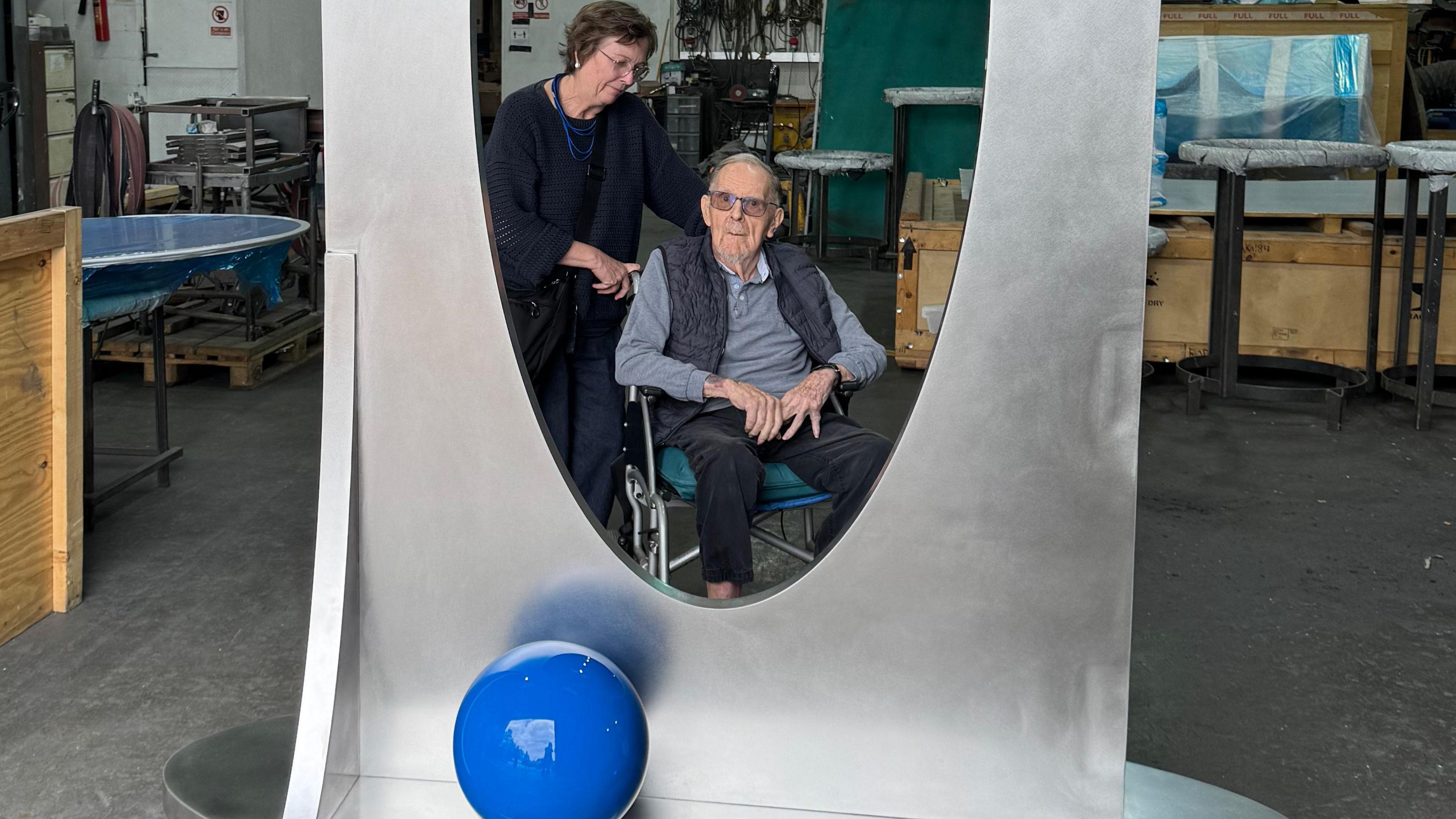 Bryan Kneale, an elderly man in a wheelchair, looking through the hole in the sculpture while it's in the workshop. His daughter Kate is standing next to him. She has short brown hair. They are both wearing glasses.