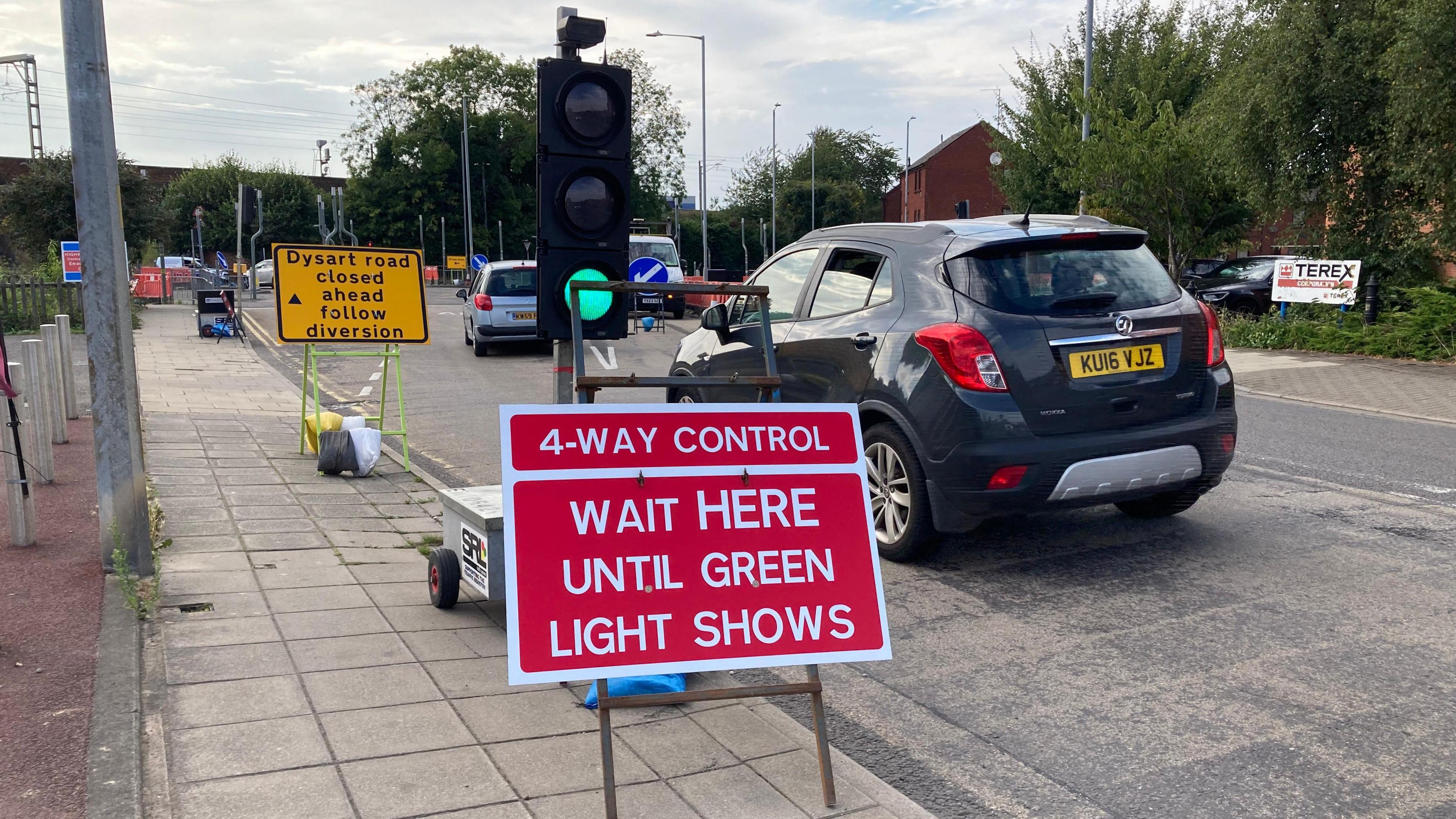 A car drives through some temporary traffic lights close to the Dysart Road closure in Grantham, there is a 'road closed' yellow sign and traffic cones and directional signs in the background.