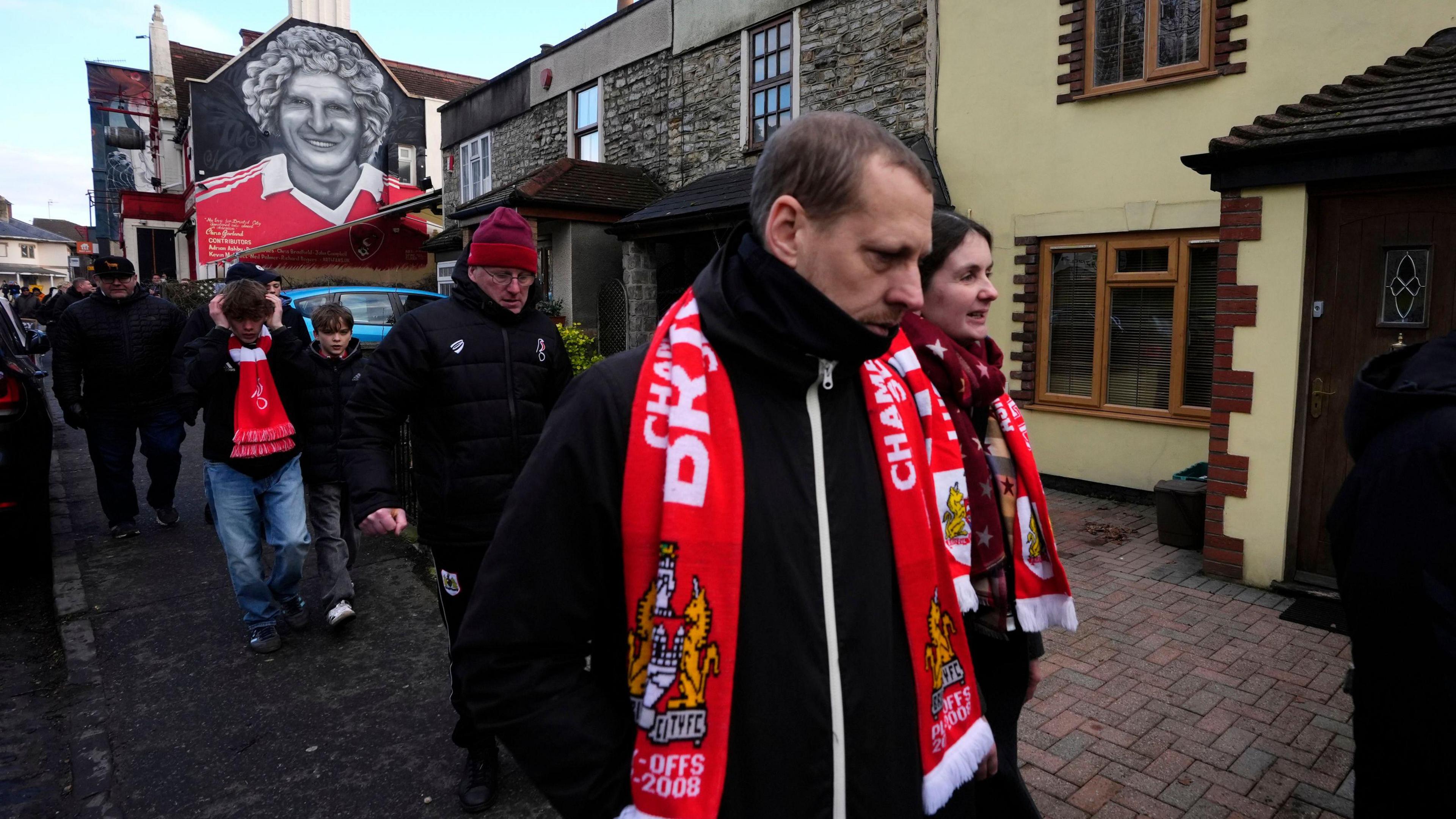 Bristol City fans, some of them wearing red and white scarves, walk towards Ashton Gate for the FA Cup tie against Wolverhampton Wanderers. Above them is a street art mural of former City player Geoff Merrick