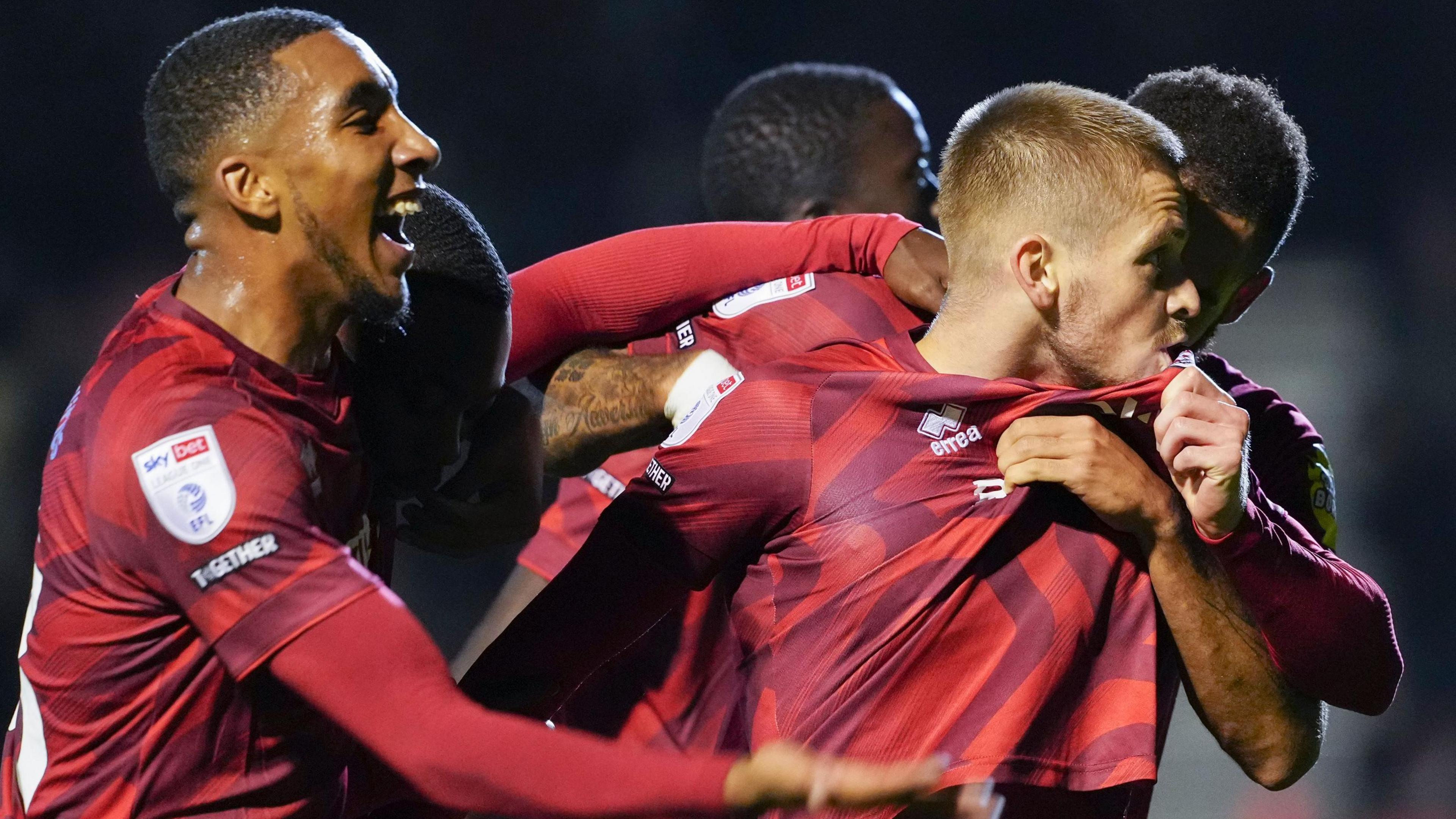 Crawley players celebrate scoring a goal against Lincoln. A player kisses the badge on his shirt while other players embrace him.