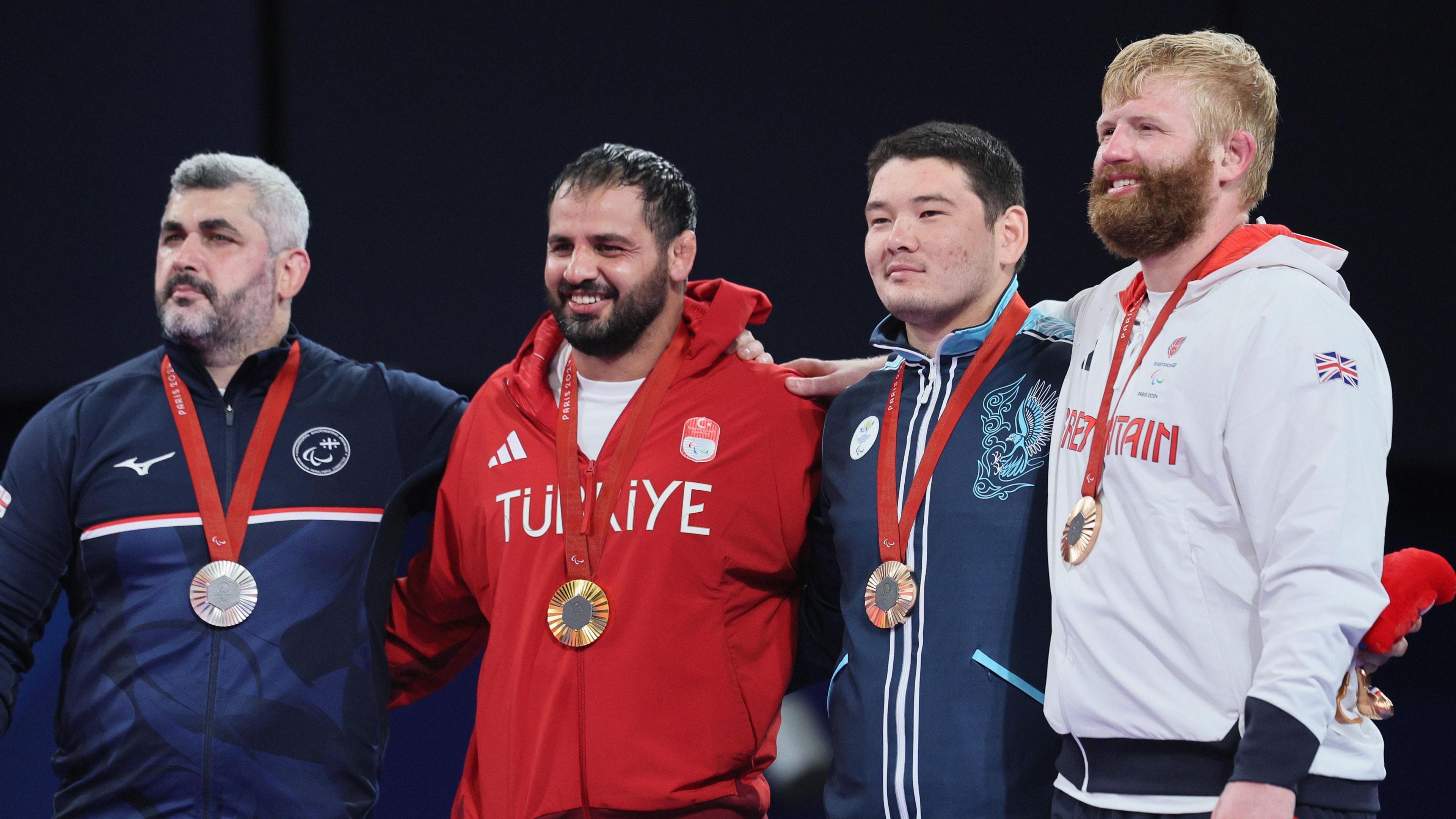 Gold medallist Ibrahim Bolukbasi of Team Turkiye (second left), silver medalist Revaz Chikoidze of Team Georgia (left), and bronze medallists Zhurkamyrza Shukurbekov of Team Kazakhstan (second right) and Christopher Skelley of Team Great Britain (right) during a medal ceremony on Day 10 of the Paris 2024 Paralympic Games. Chris Skelley is in white kit, wearing his bronze medal, and he has a thick ginger beard