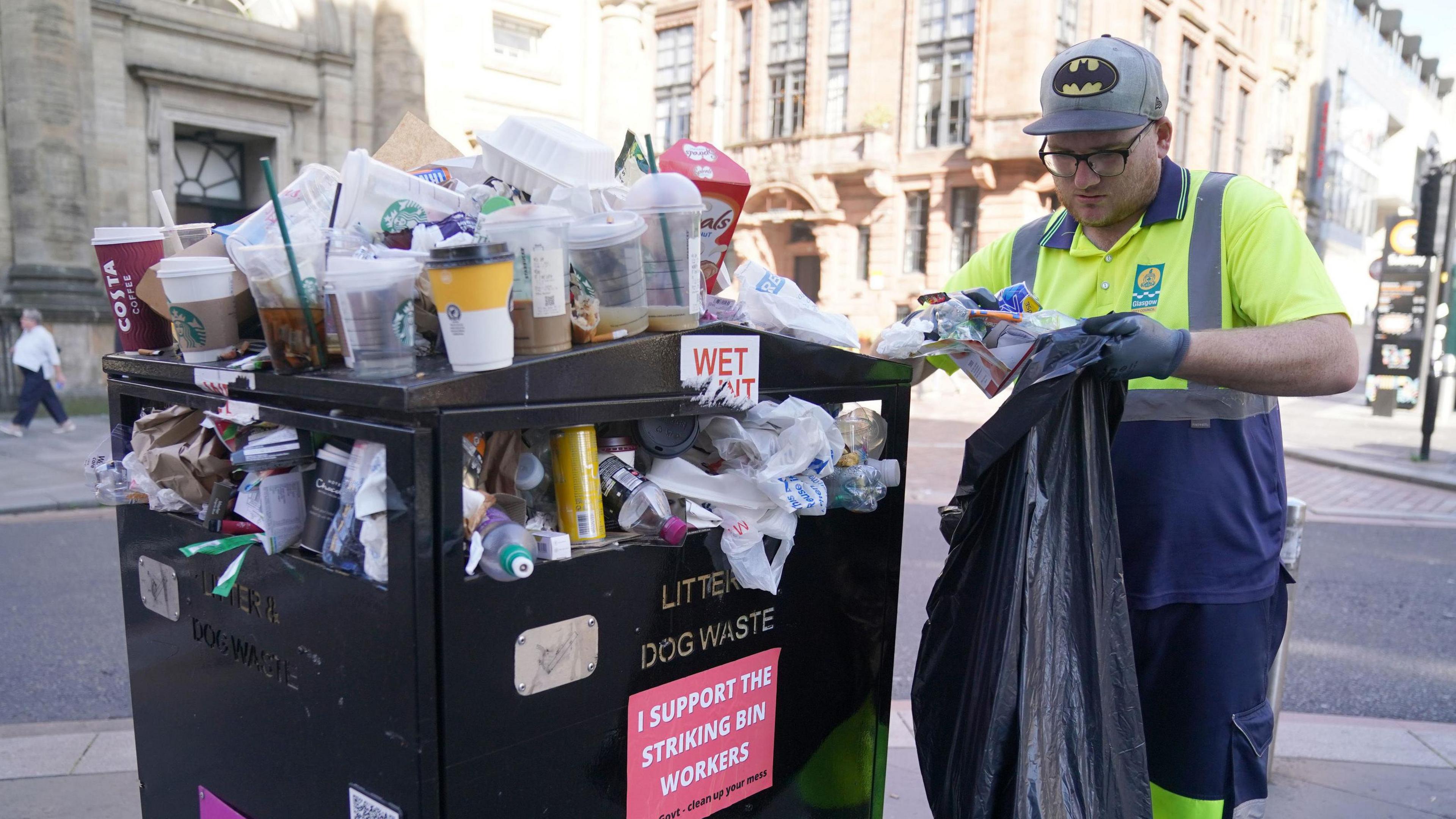 An overflowing bin next to a man in a Glasgow City Council uniform, holding a black bin bag