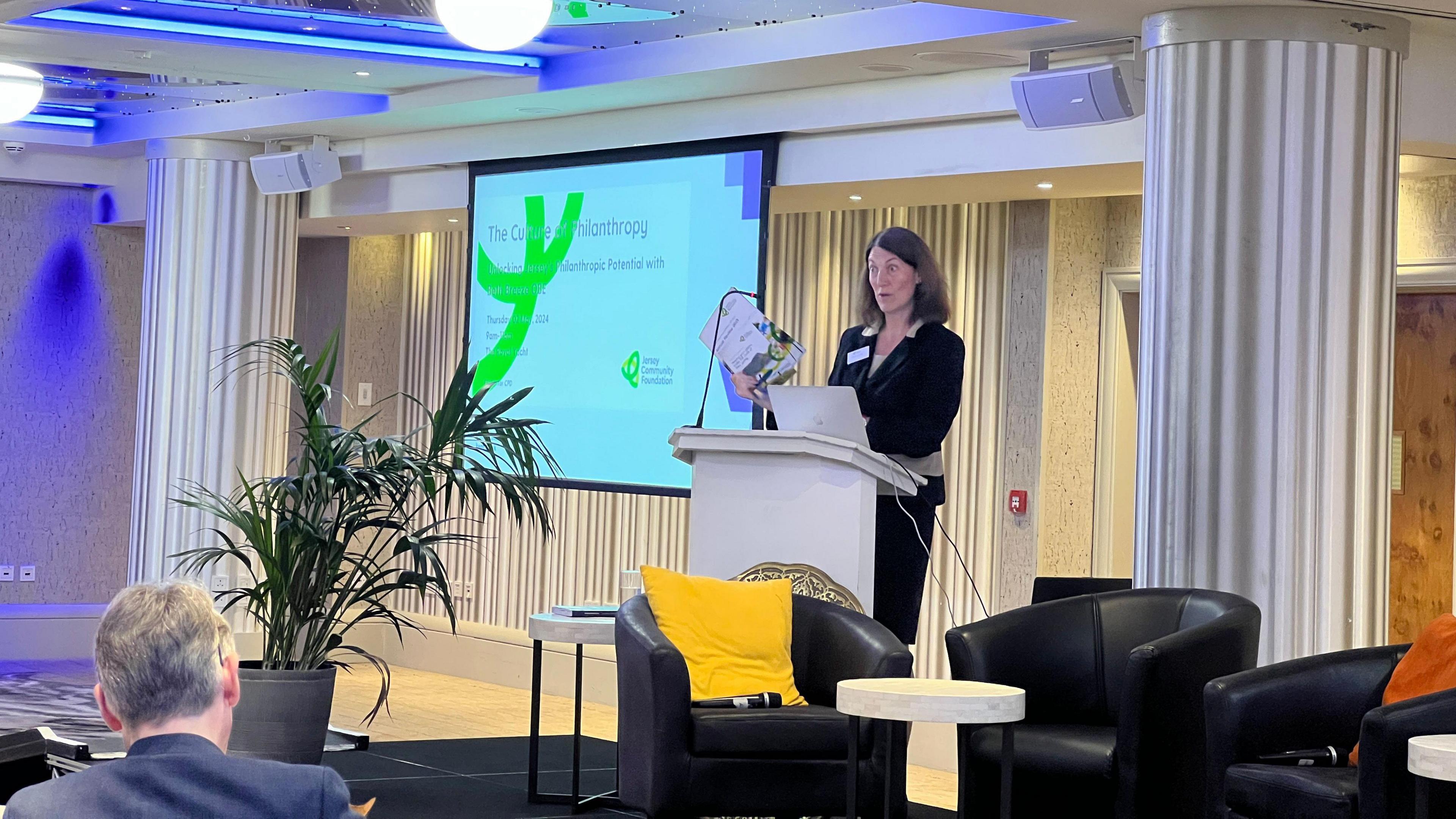 Prof Beth Breeze gives a speech on a podium holding a leaflet as a point power presentation is displayed behind her