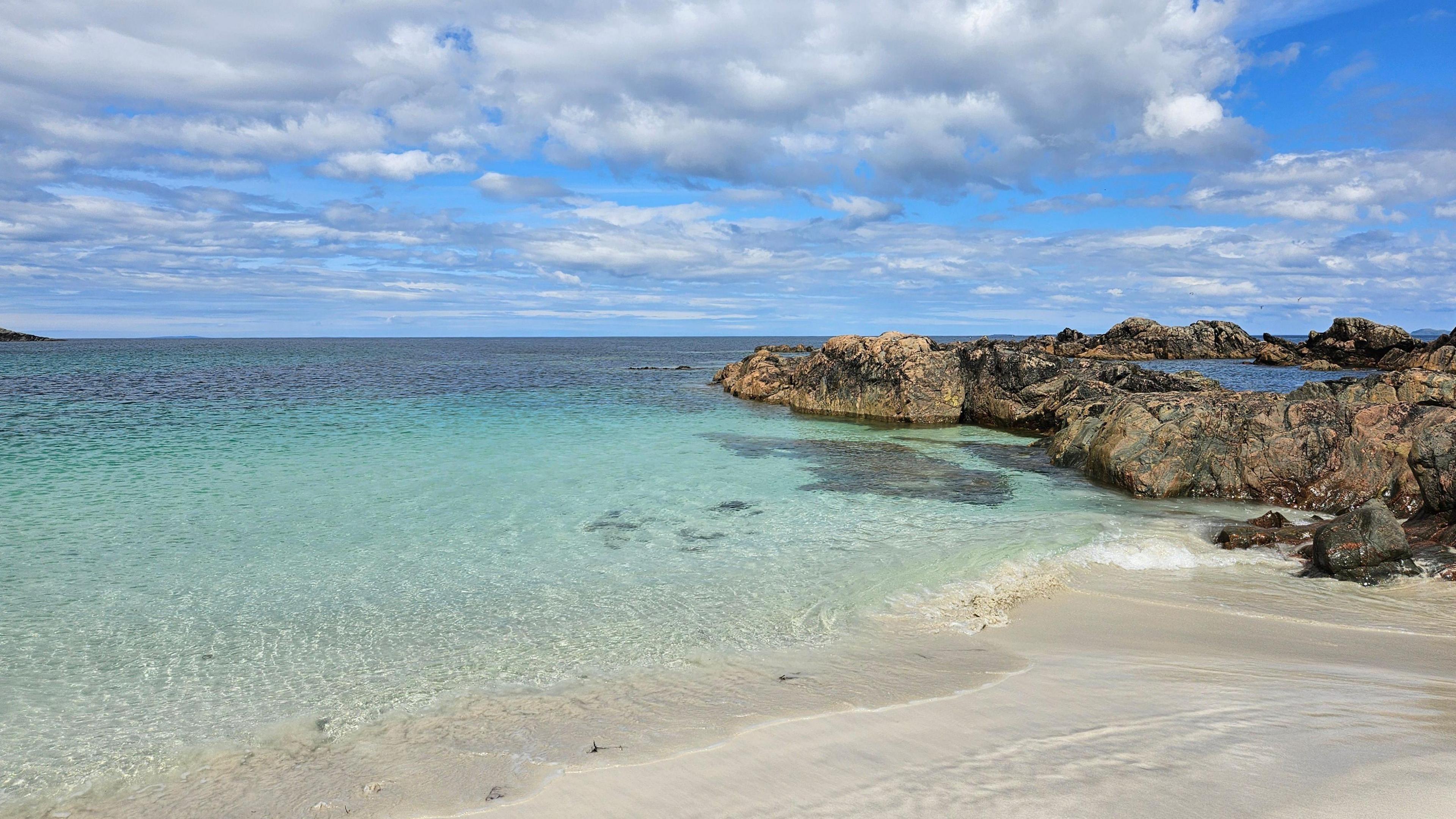 A light-colored sandy beach is in the foreground with clear, shallow turquoise water coming onto the shore. Dark-coloured rocks are to the right of the image between the sand and the water. The background displays a bright blue sky with fluffy white clouds.