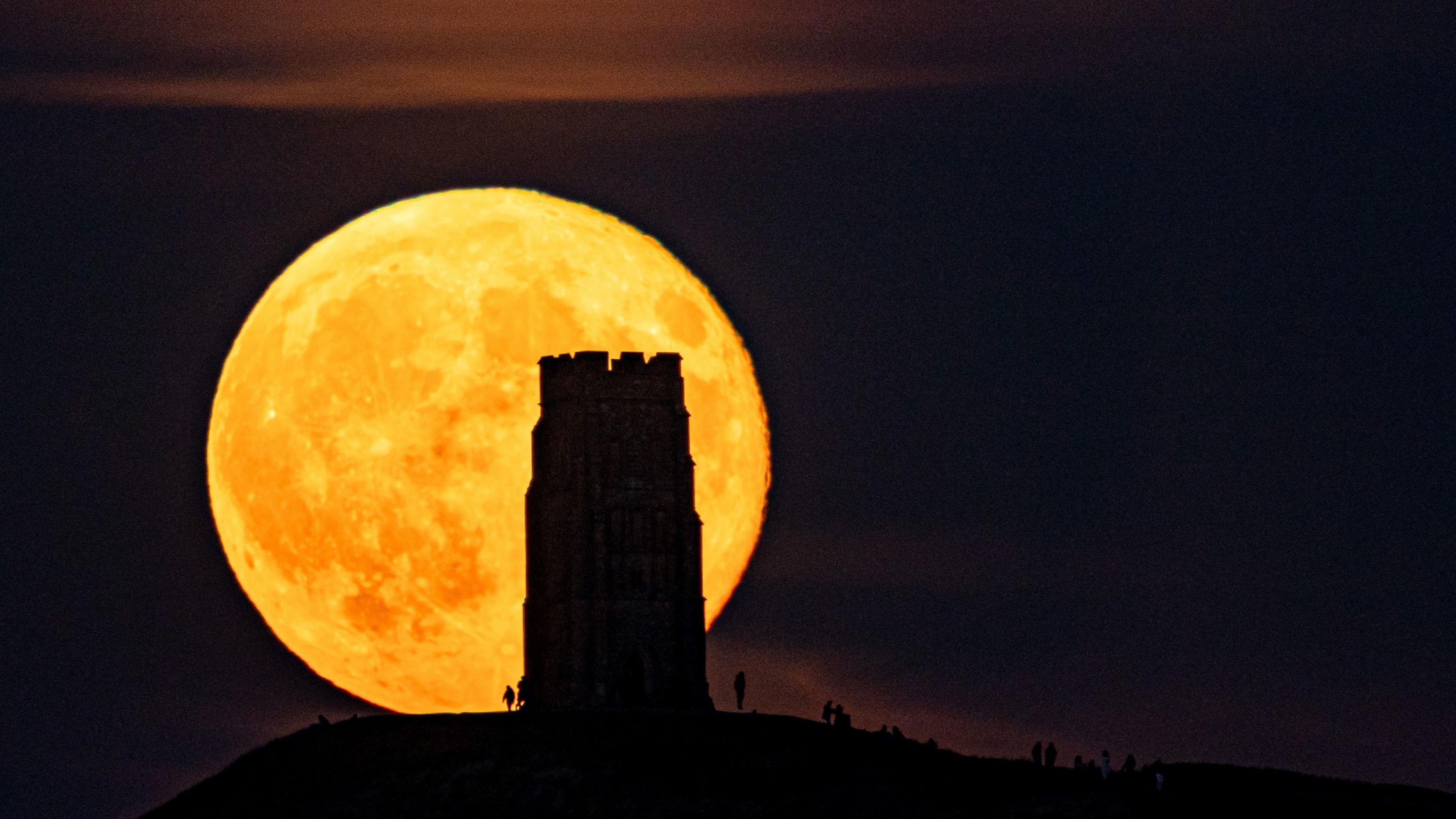 Large, orange, illuminated supermoon, rising behind a silhouetted St Michael's Tower in Glastonbury with people gathering in the distance under a dark night sky